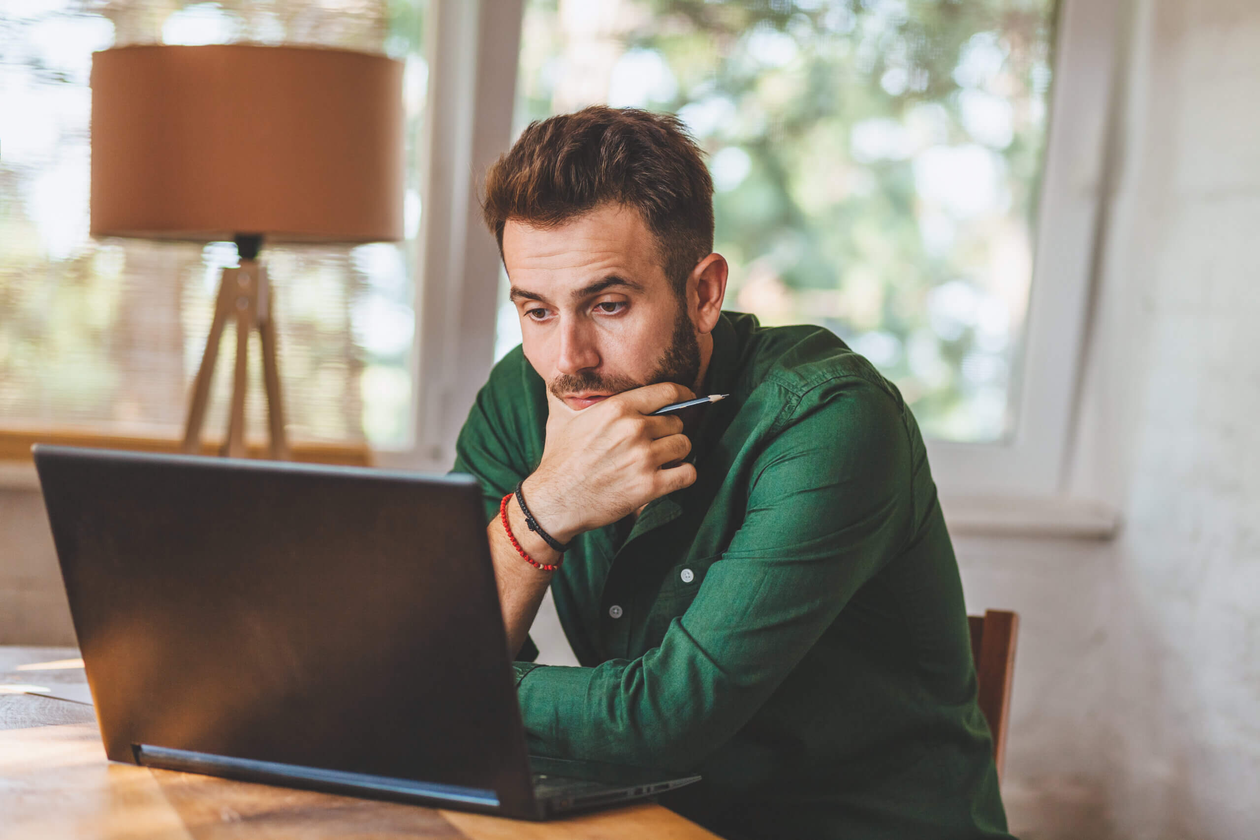A man in a green shirt sits at a table, looking intently at a laptop screen. He holds a pen in his right hand and rests his chin on his left hand. A lamp and window with blurred foliage are in the background.