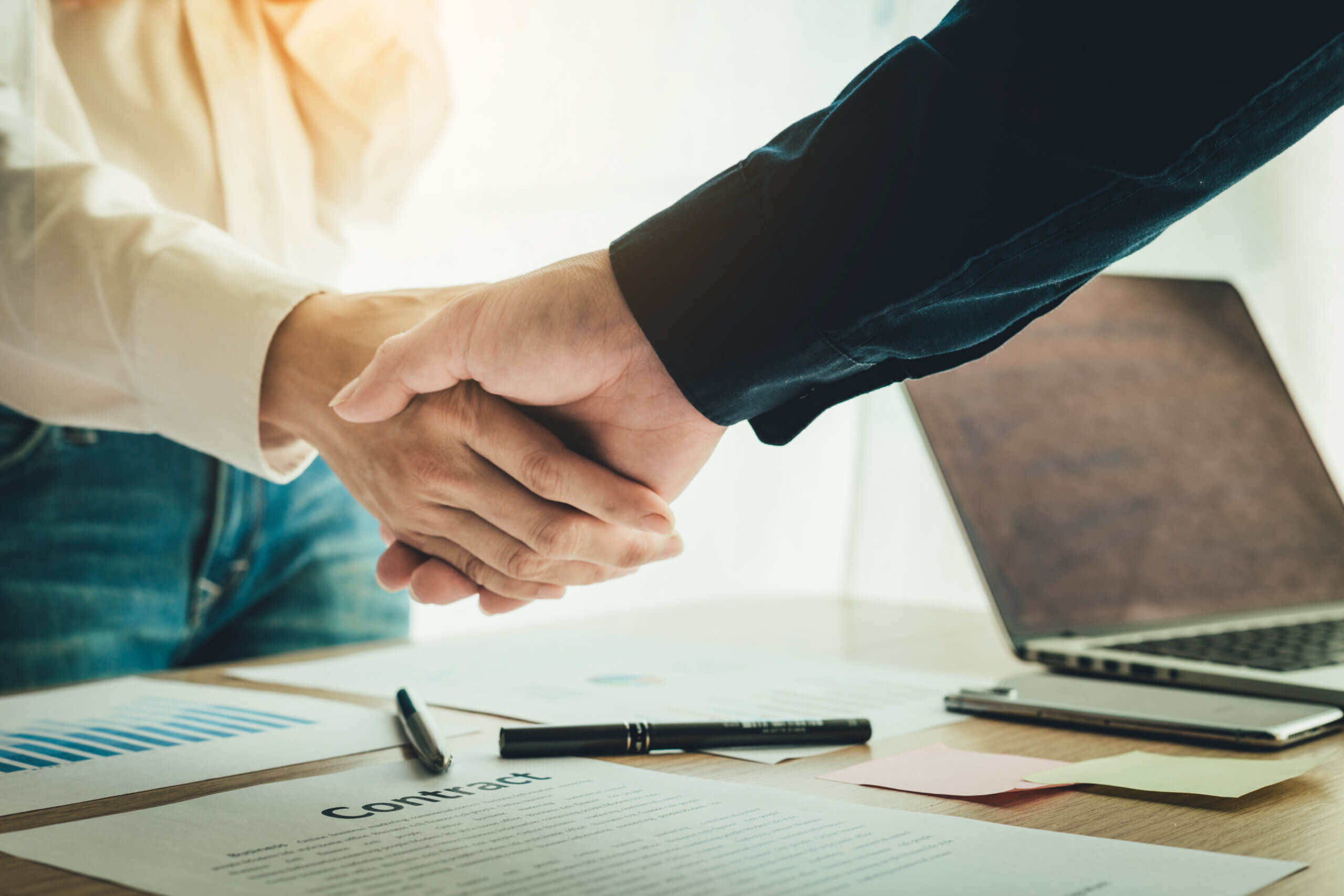 Two people shaking hands over a wooden table with paperwork, including a document labeled "Contract," graphs, and a pen. A laptop and sticky notes are visible, indicating a business agreement or partnership setting.