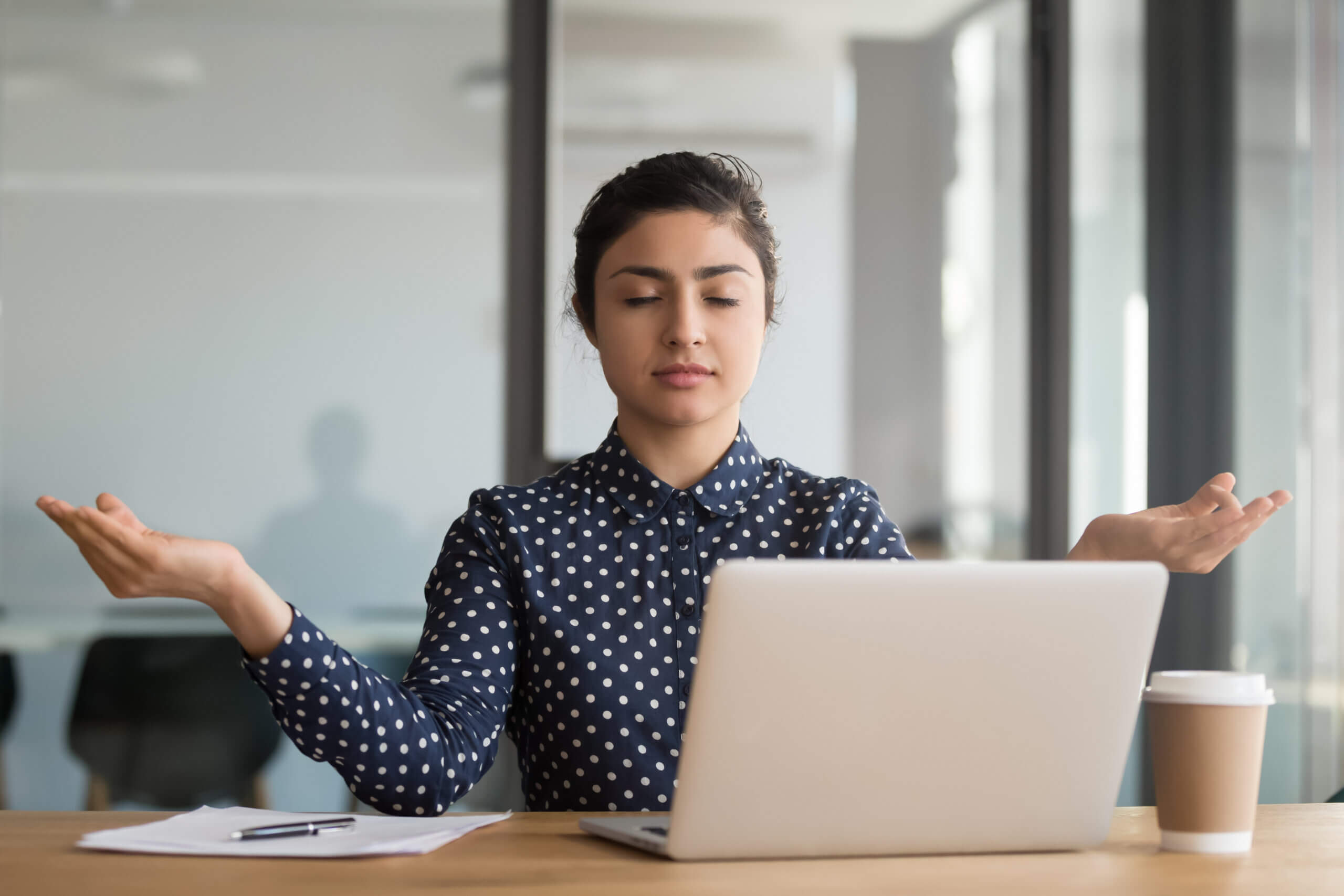 A person sits at a desk with a laptop, eyes closed, and arms raised in a calm, meditative pose. Papers and a coffee cup are on the desk. They are wearing a dark polka-dotted shirt. The setting appears to be a modern office with glass walls.