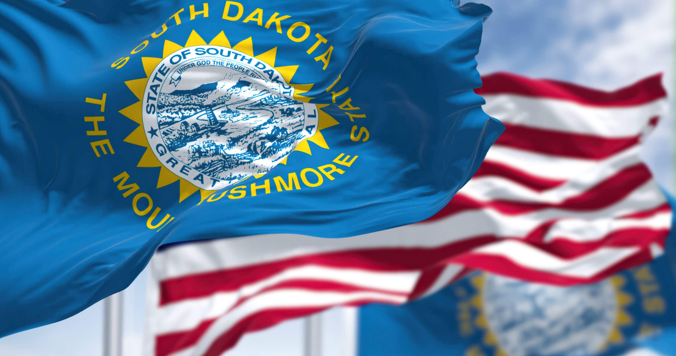 The image shows the South Dakota state flag prominently in the foreground, featuring a blue field with the state seal surrounded by a yellow sun and the words "South Dakota" and "The Mount Rushmore State." American flags wave in the blurred background.