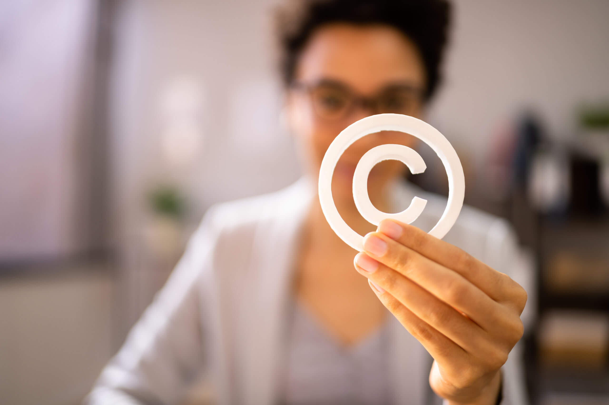 A person holding a white copyright symbol in focus, with their face blurred in the background. They have short curly hair and are wearing glasses and a gray blazer. The background is softly lit and out of focus.
