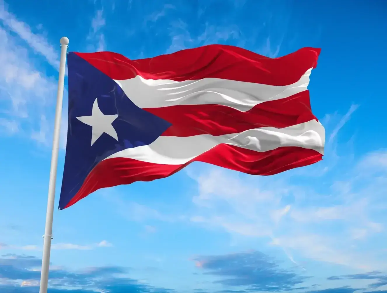 The Puerto Rican flag with red and white stripes and a white star on a blue triangle waves against a bright blue sky.