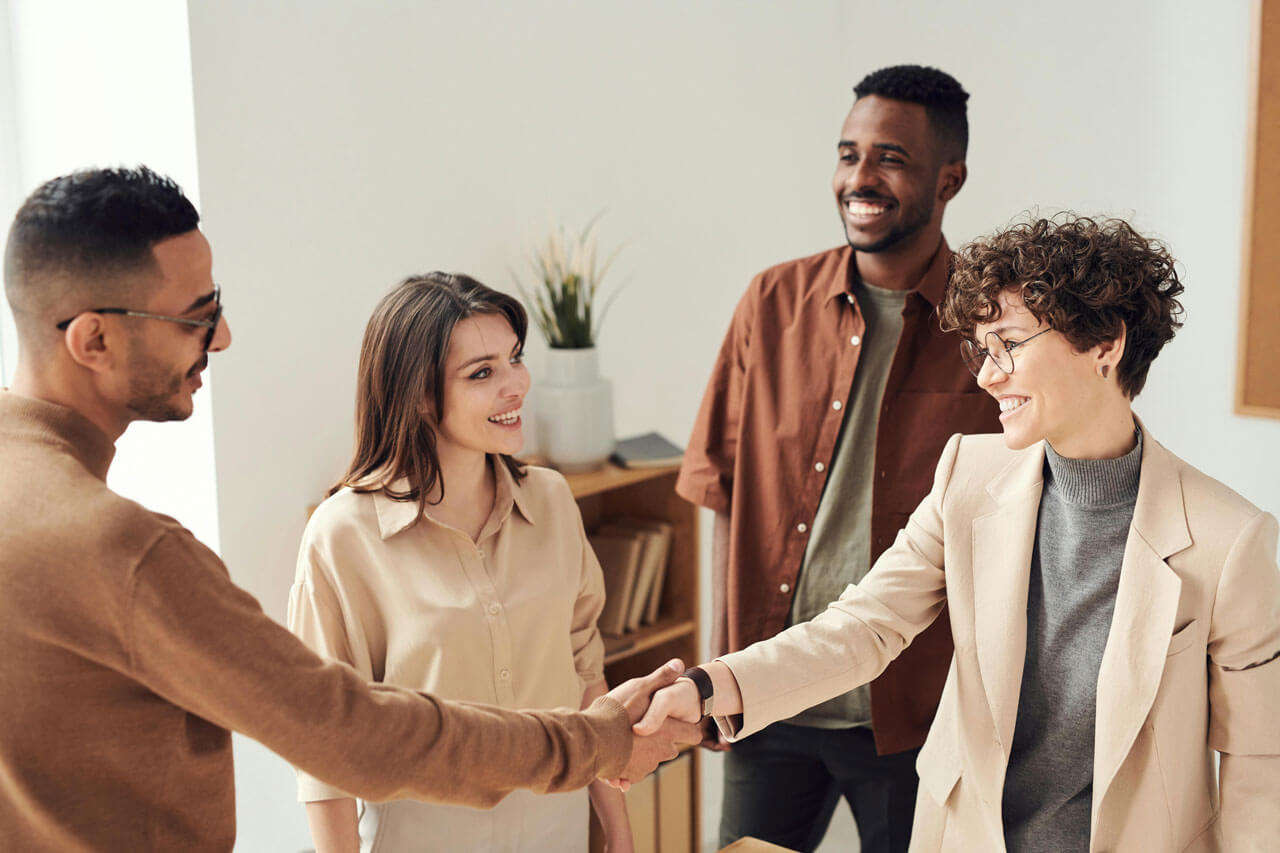 Four people stand in an office setting. Two are shaking hands while smiling, and the other two are smiling and looking on. One person wears glasses. A plant and shelves are visible in the background.
