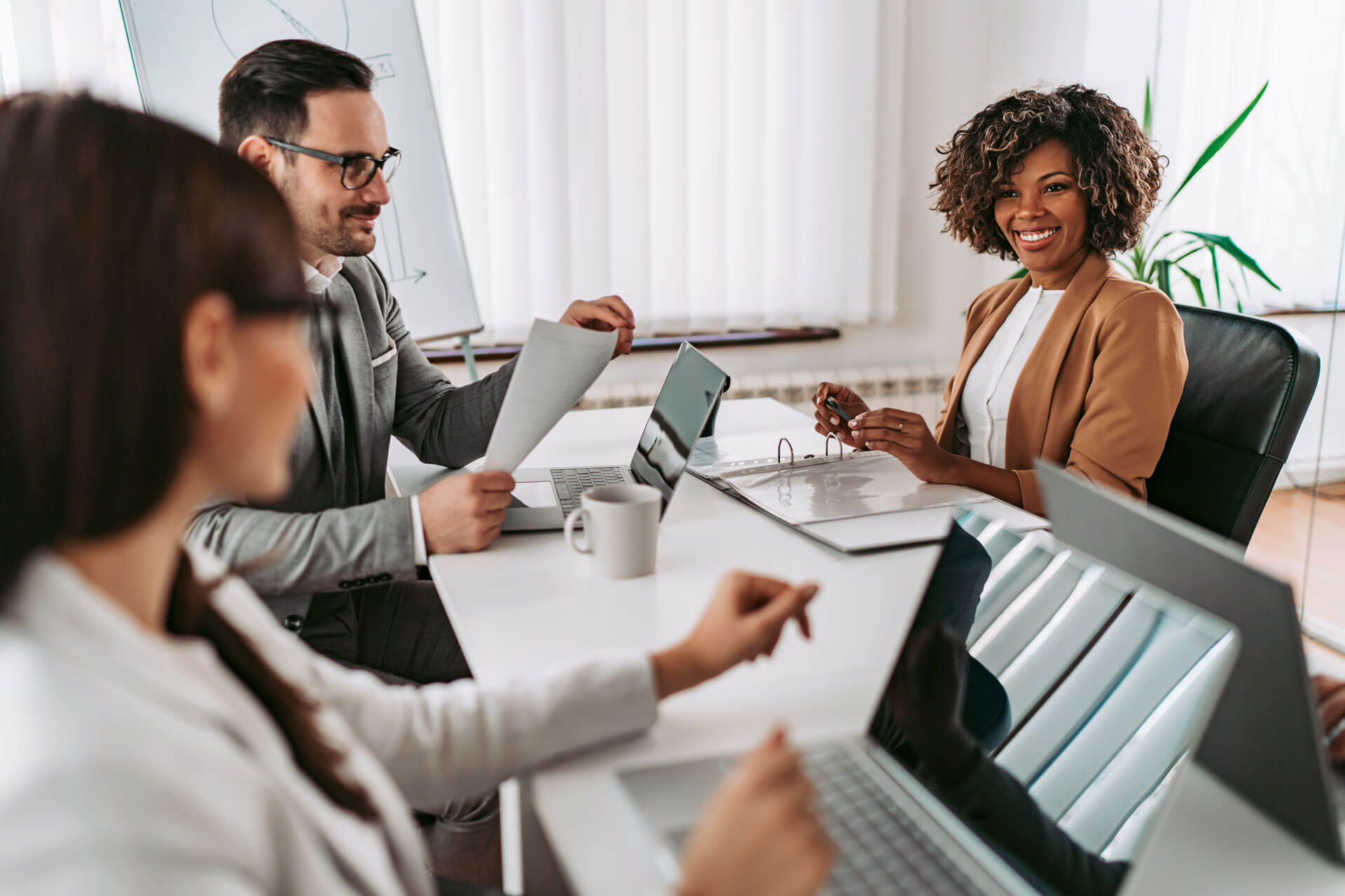 Three professionals in a meeting room engaged in discussion around a table with laptops, papers, and a coffee cup. One person is smiling while another holds a document. A whiteboard and a window with blinds are in the background, representing Cogency Global's registered agent services and a collaborative business environment.