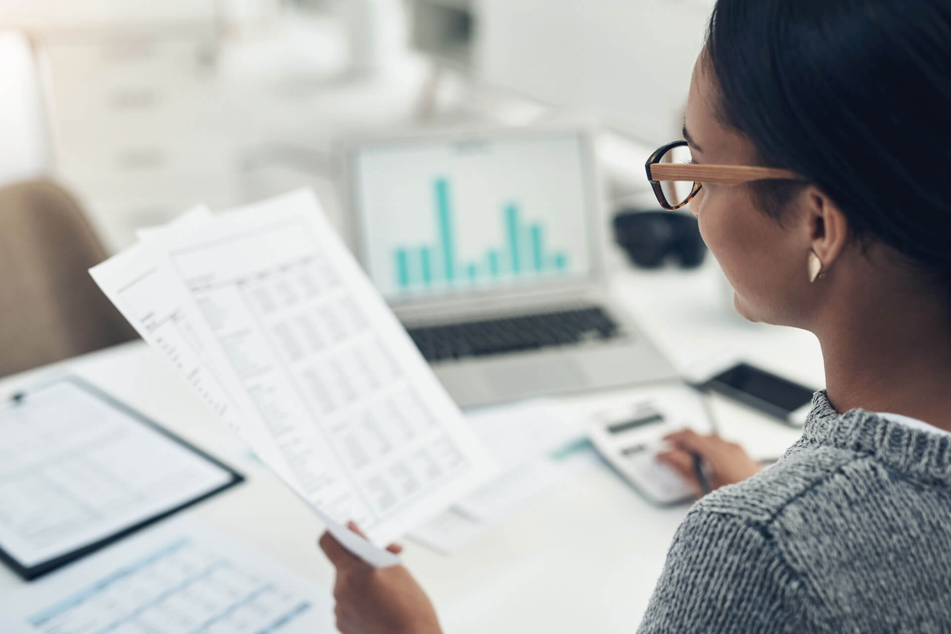 A person wearing glasses is seated at a desk, holding documents and working on a laptop. A calculator, other papers, and office supplies are visible on the desk. A bar graph is displayed on the laptop’s screen, illustrating data analysis or financial work.