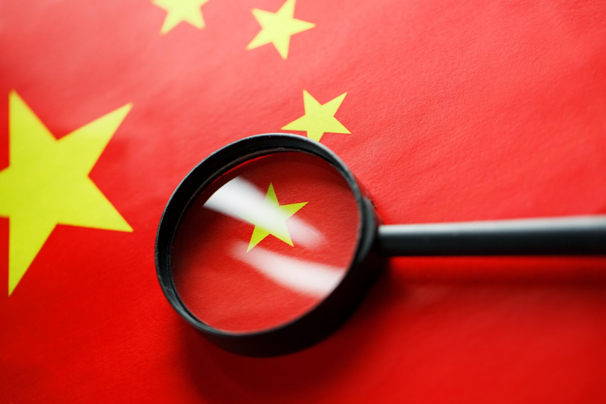 A magnifying glass placed over a small star on the Chinese flag, emphasizing its details. The flag is red with one large star and four smaller stars, all yellow, located in the top left corner.