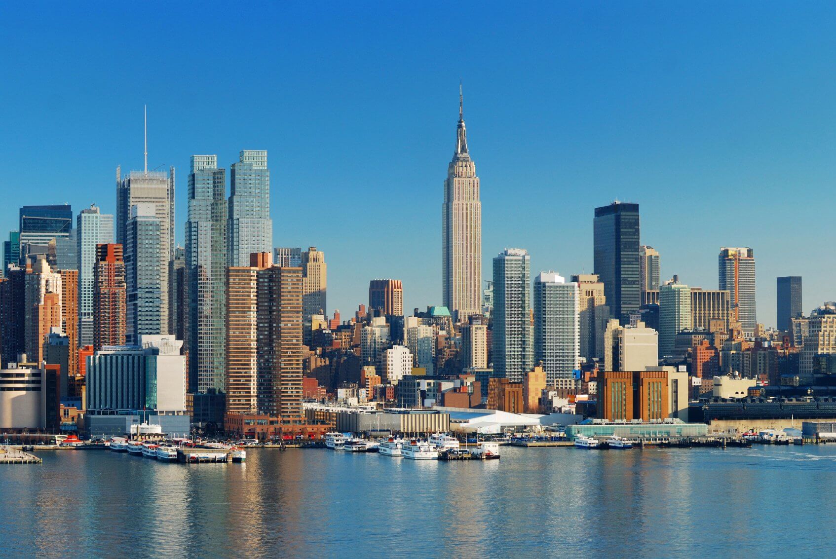 A city skyline view of New York City featuring the Empire State Building and surrounding skyscrapers under a clear blue sky, with waterfront buildings and boats in the foreground reflecting on the water.