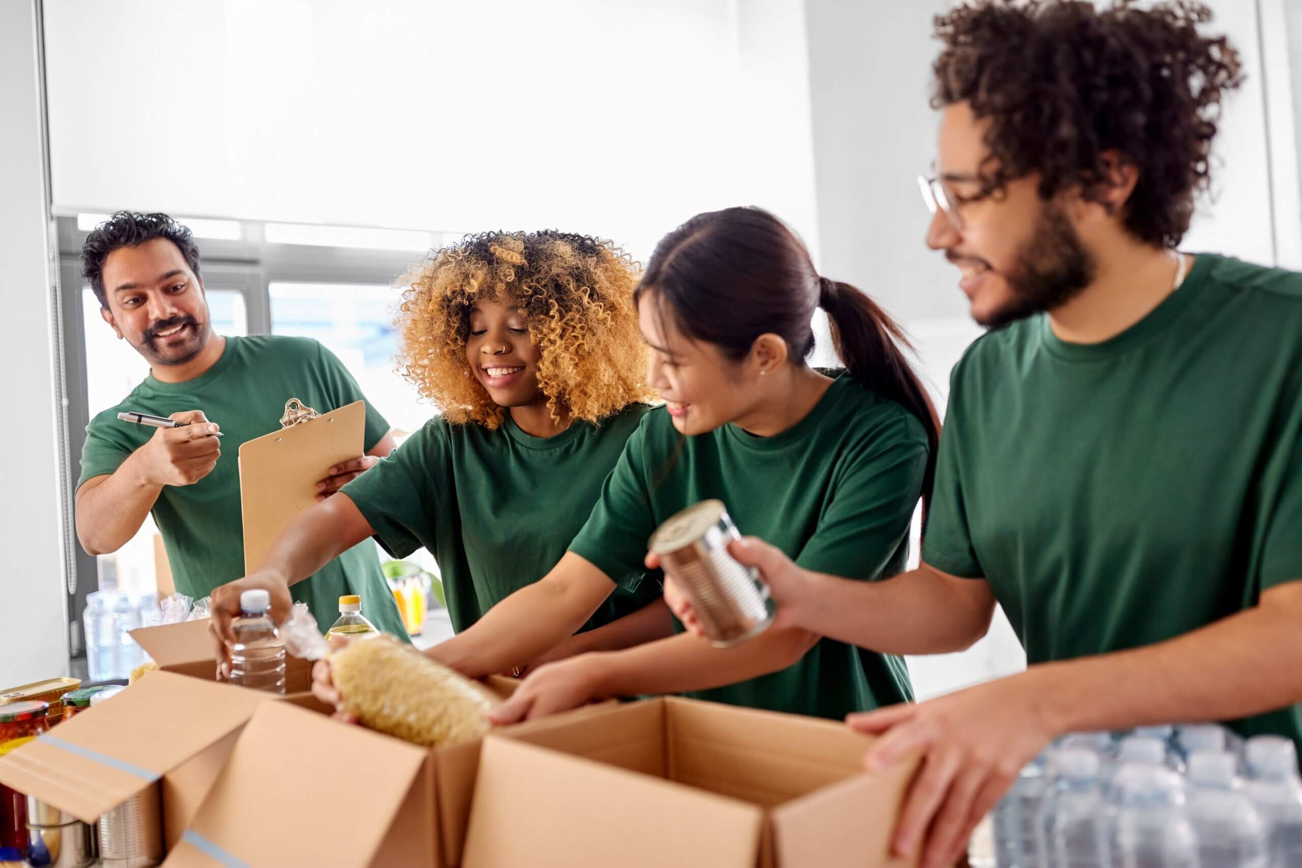 Four people in green shirts are packing food items into cardboard boxes. They are smiling and focused on their task. One person is holding a clipboard, while the others handle canned goods and pasta. Water bottles are on the table.