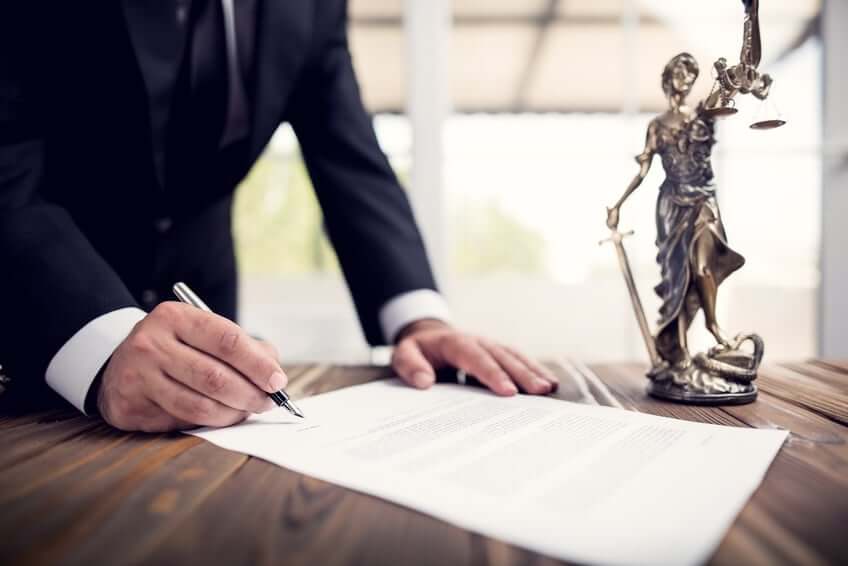 A person in a suit signs a document on a wooden table. Beside the document is a bronze statue of Lady Justice holding scales and a sword. The background is softly blurred with bright light coming through windows.