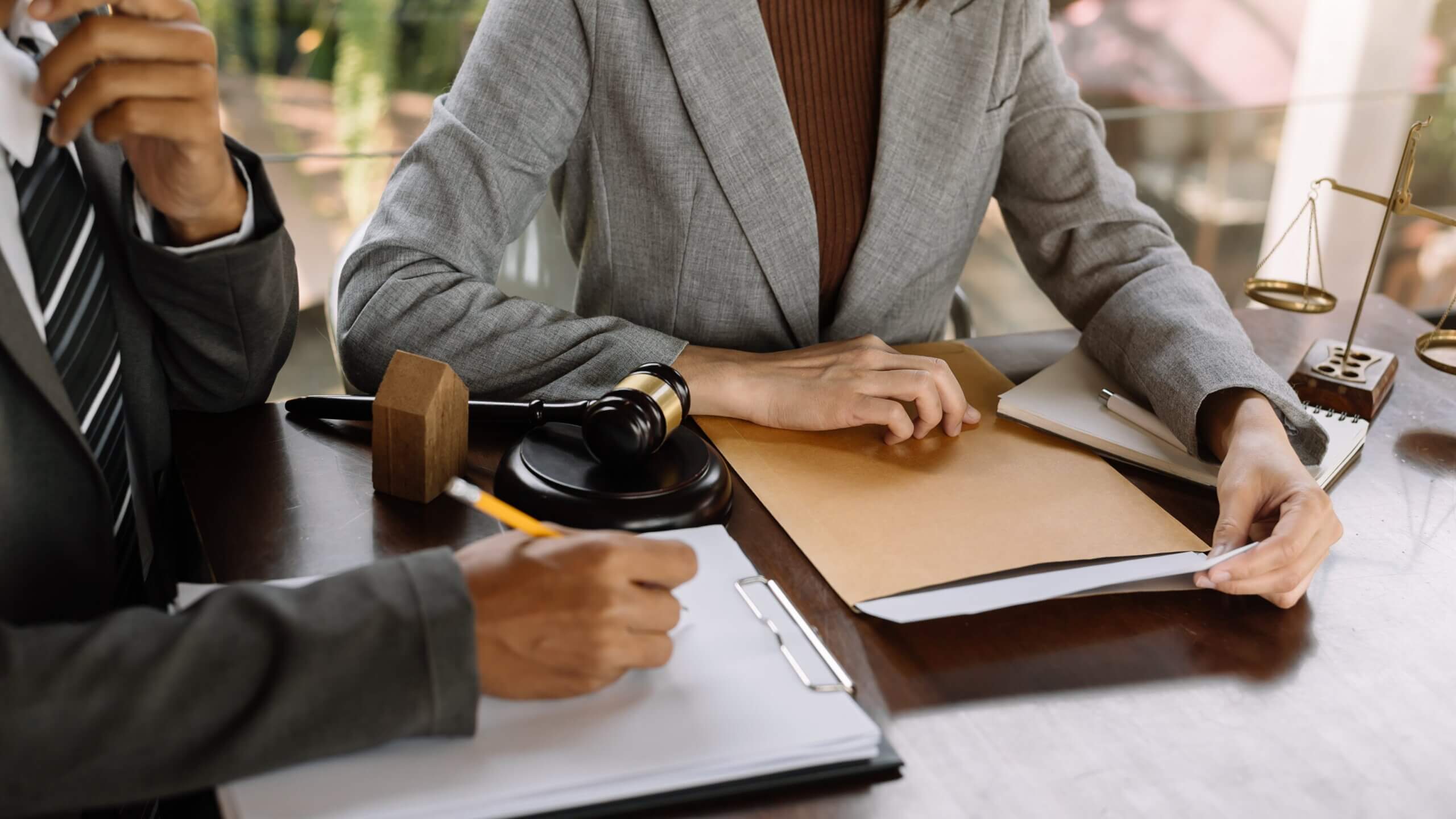 Two professionals sit at a table in an office setting, wearing suits. One is holding a file, while the other writes on a clipboard. A gavel and scales of justice are on the table, suggesting a legal consultation.