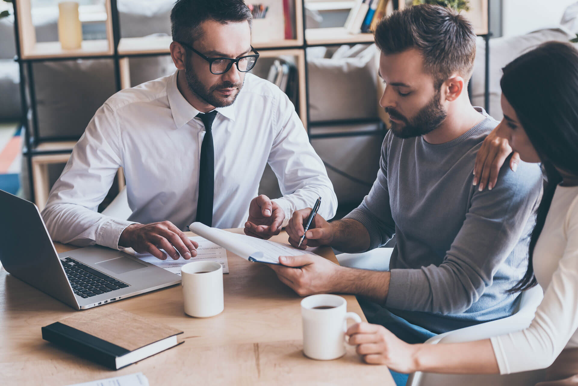 A man in a dress shirt and tie is speaking with a couple who are signing a document, representing Cogency Global’s charitable solicitation registration and renewals service. A laptop and two coffee mugs are on the table.