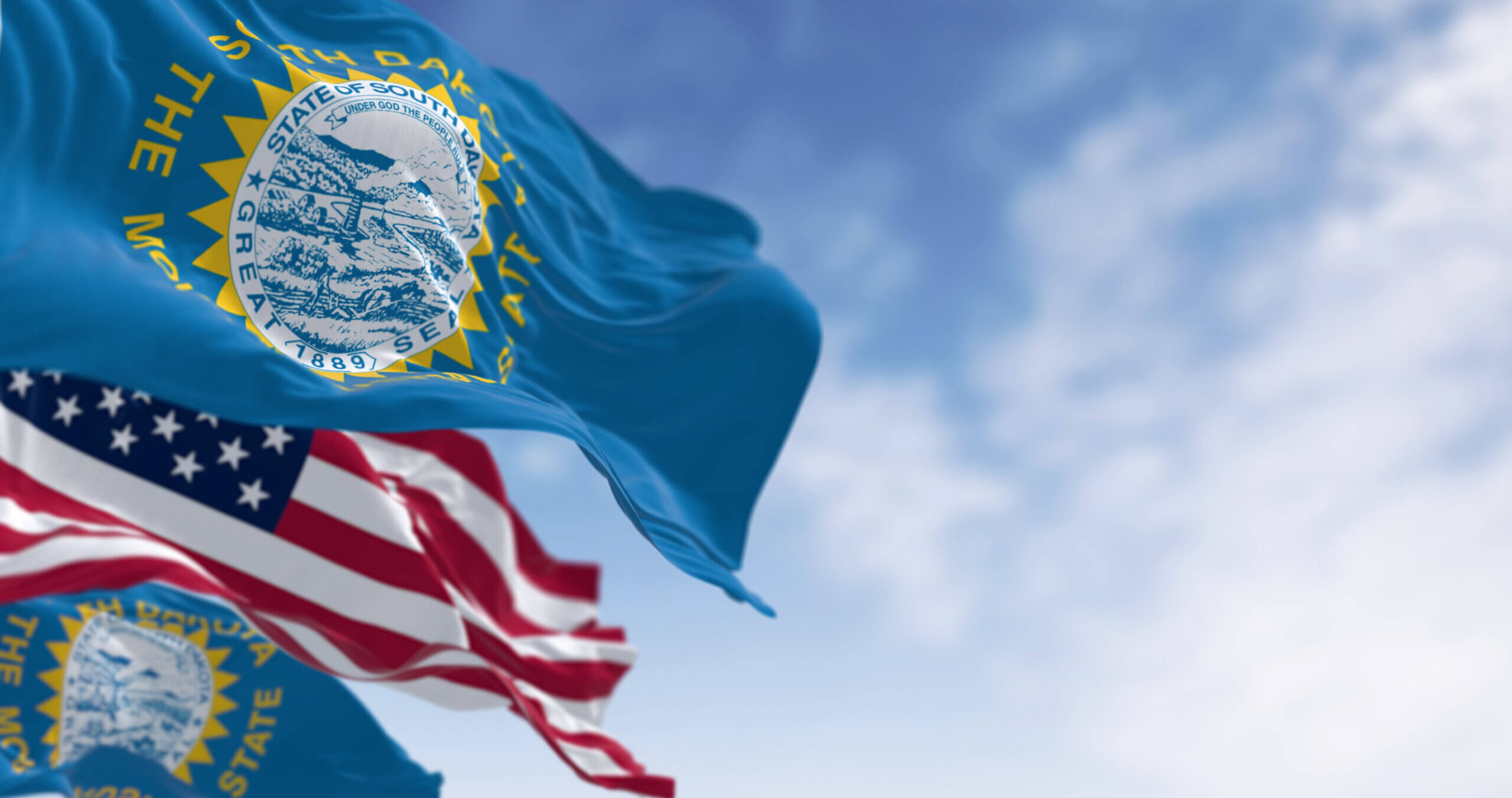 Two blue South Dakota state flags and one United States flag wave in the wind against a blue sky with scattered clouds.