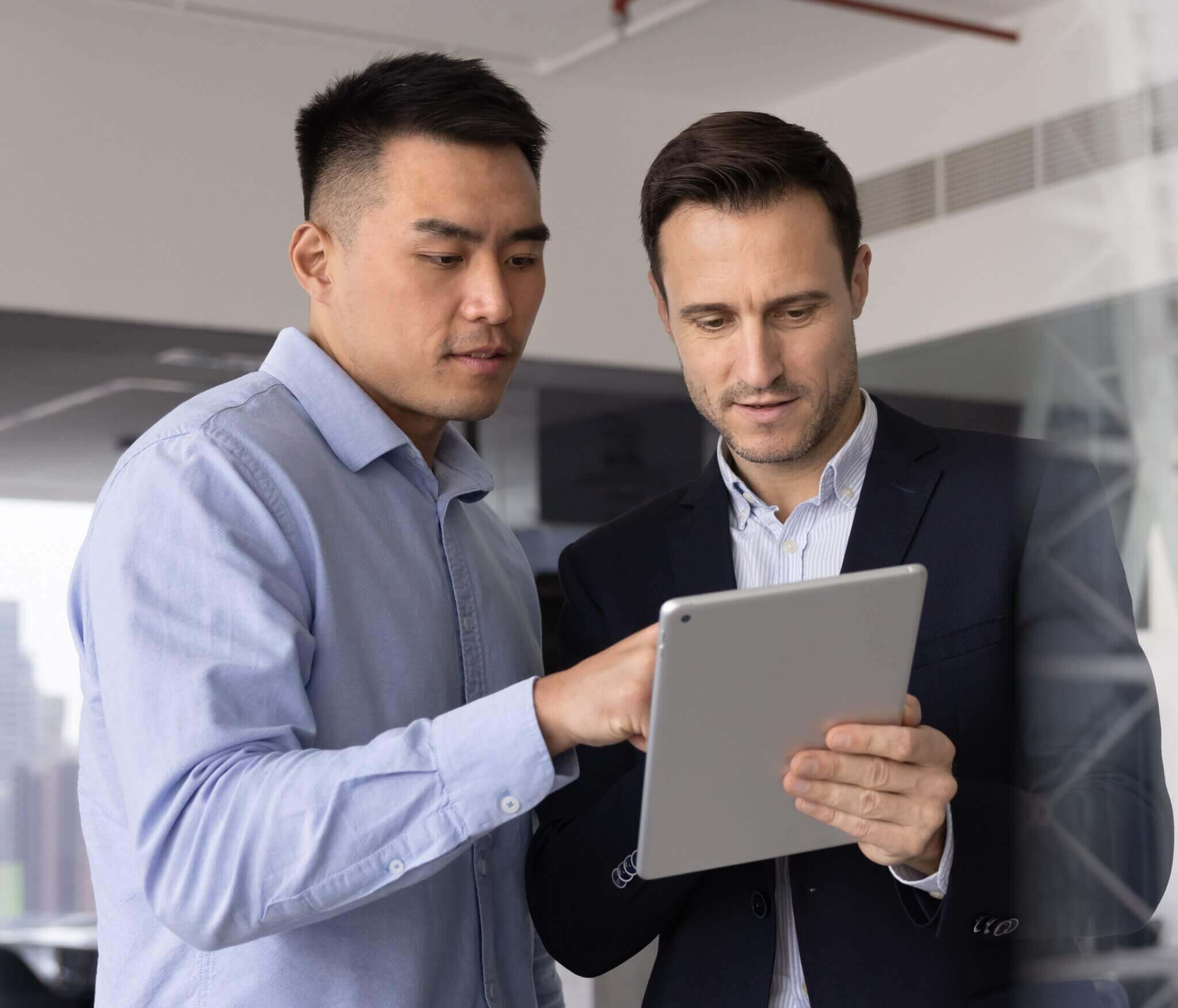 Two men in business attire looking at a tablet together in an office.