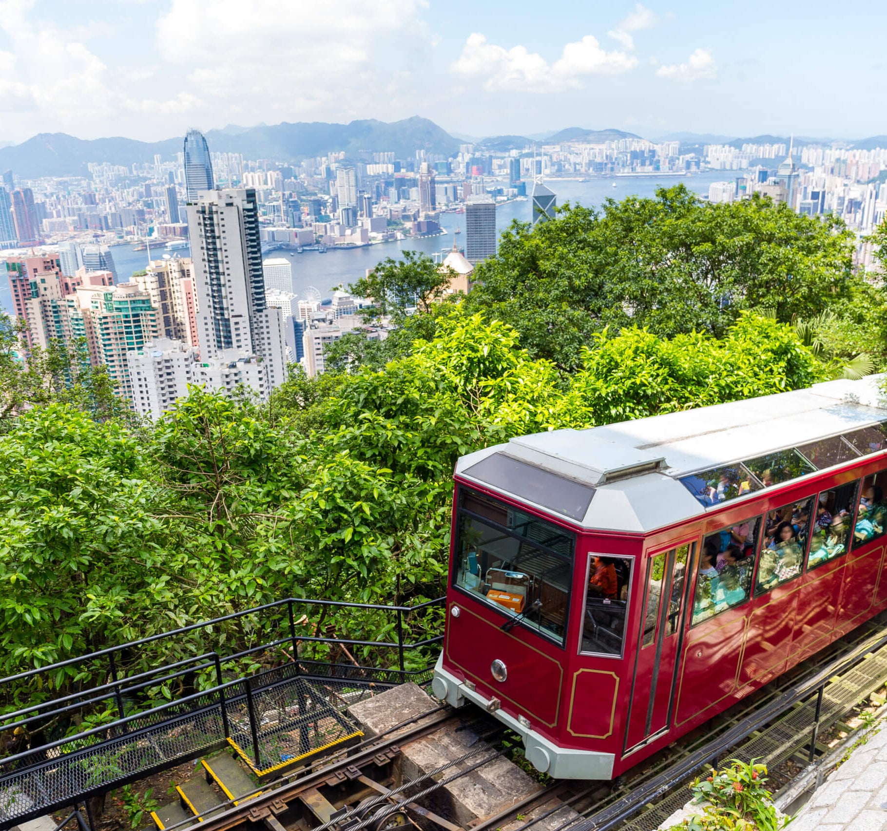 A red Peak Tram ascending a steep track with a view of the Hong Kong skyline in the background.