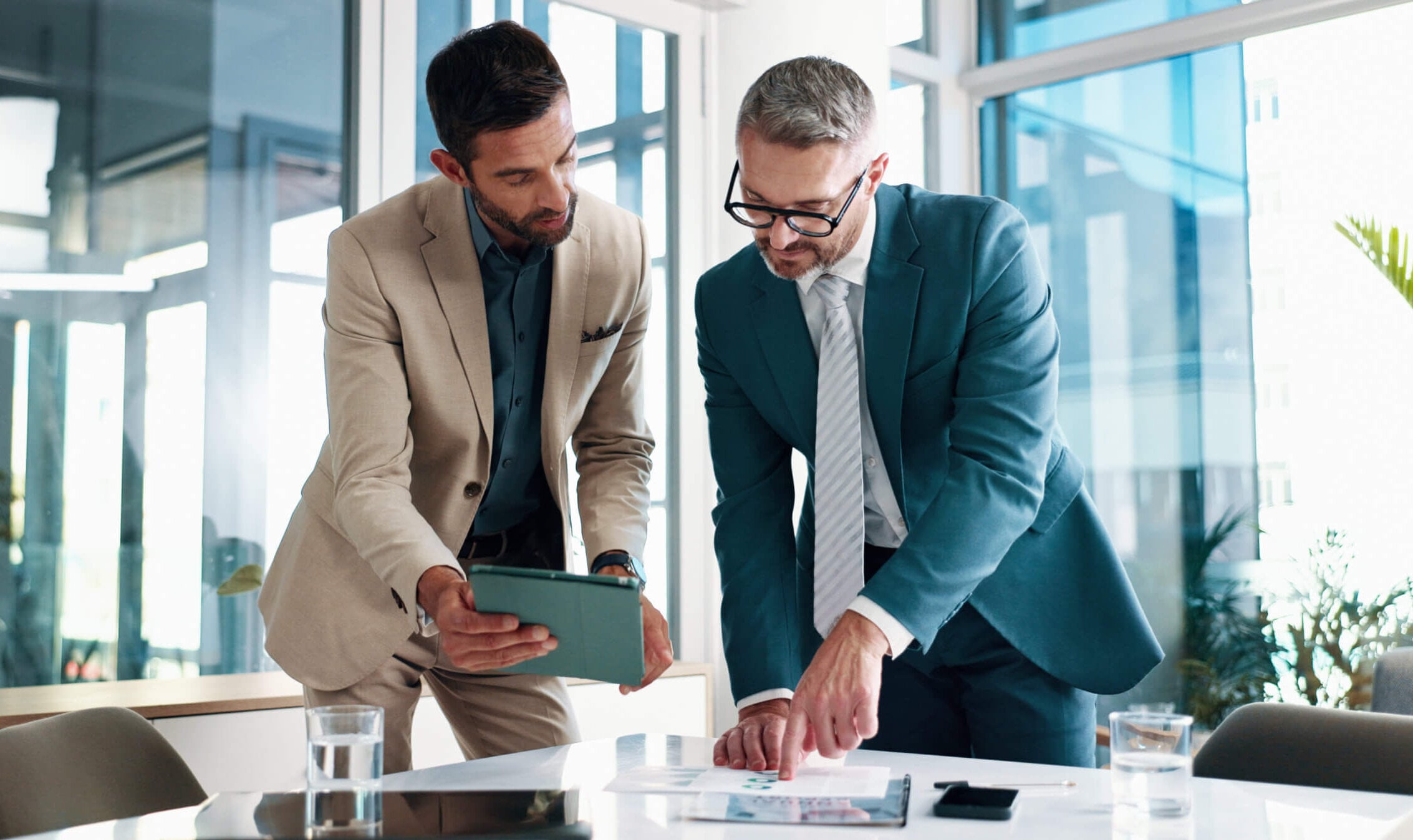 Two men in suits stand at a table in an office, one holding a tablet and the other pointing at papers. They appear to be discussing business documents.