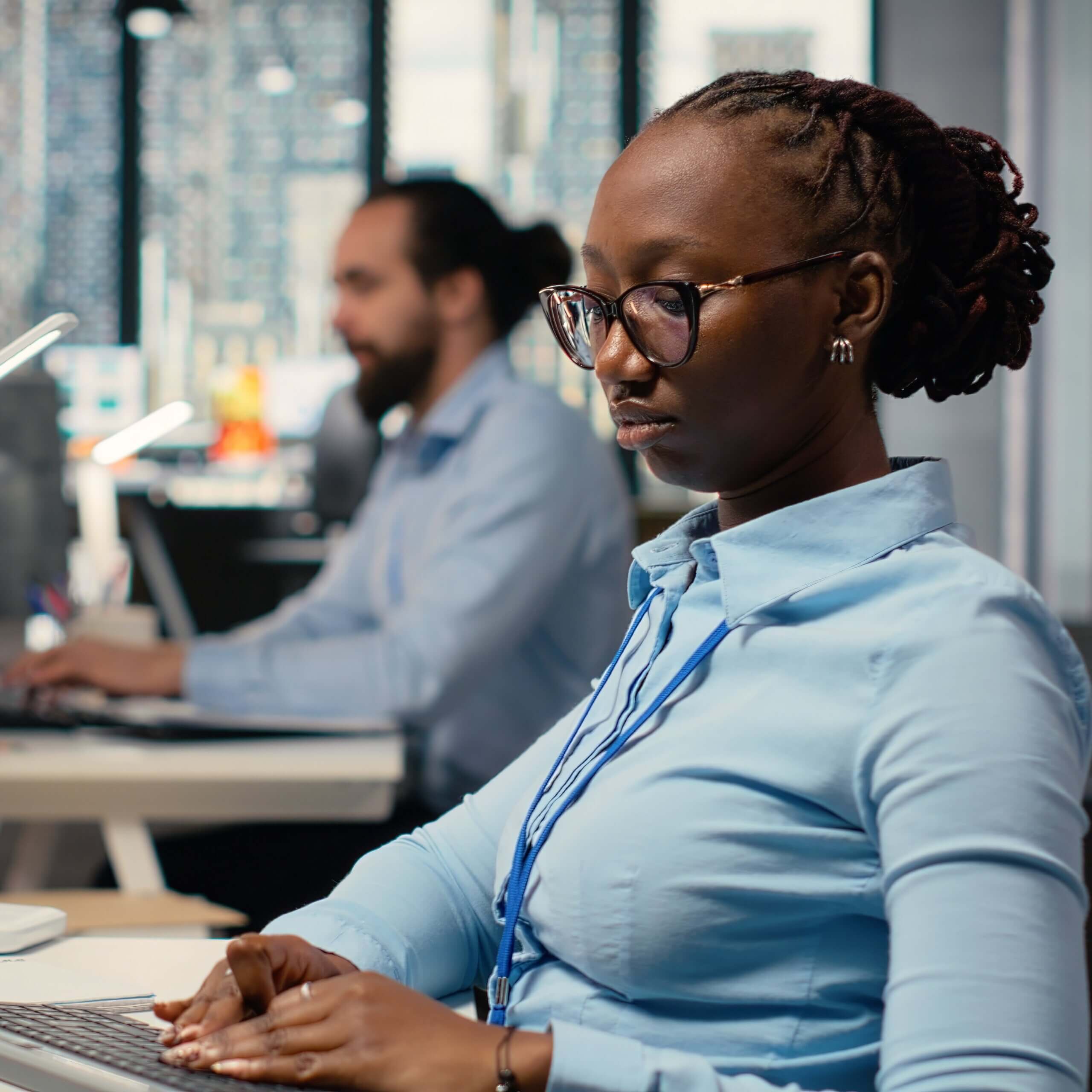 A woman in glasses and a blue shirt works on a computer at an office desk, with a man working at another desk in the background. The office has large windows and a modern, professional atmosphere.
