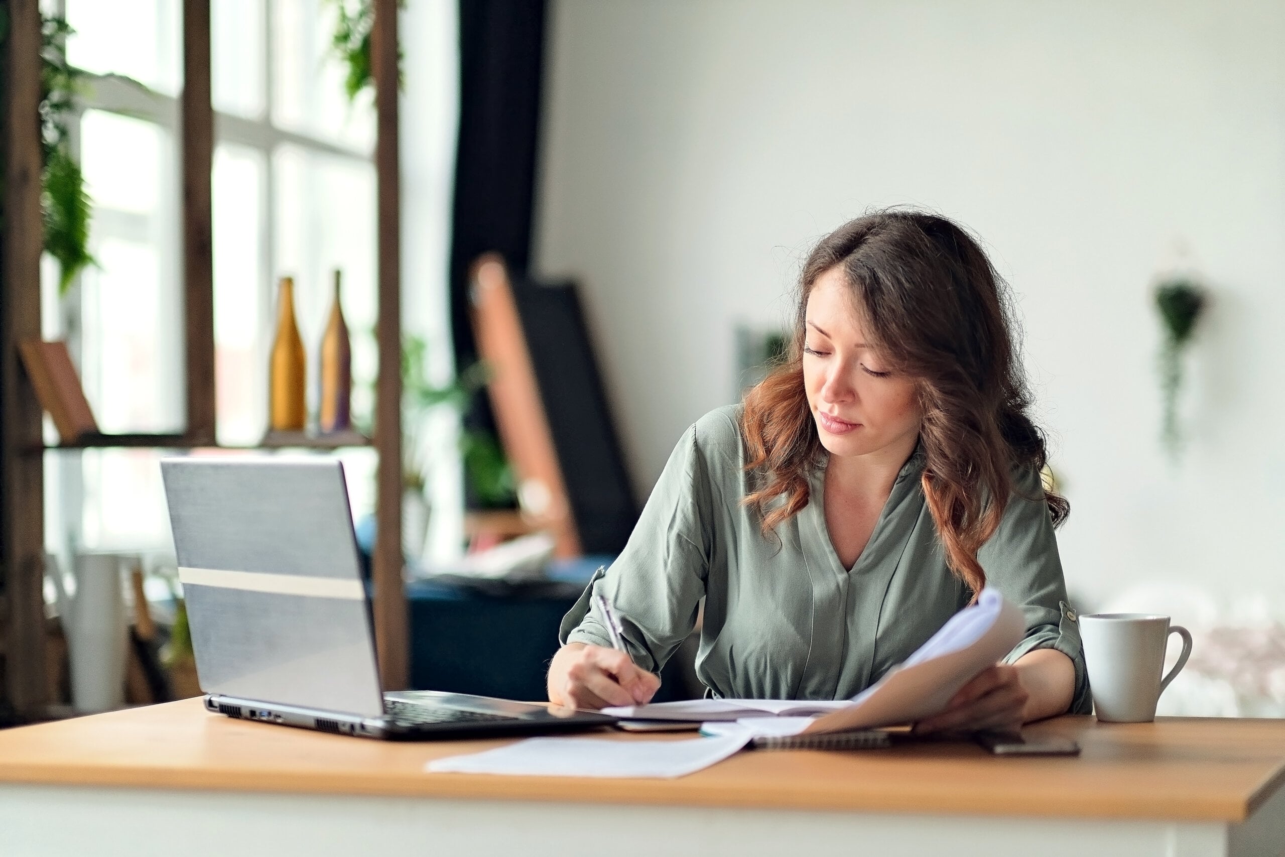 Woman with wavy hair sits at a desk, writing in a notebook beside an open laptop and a white mug, in a bright room with large windows and plants in the background.