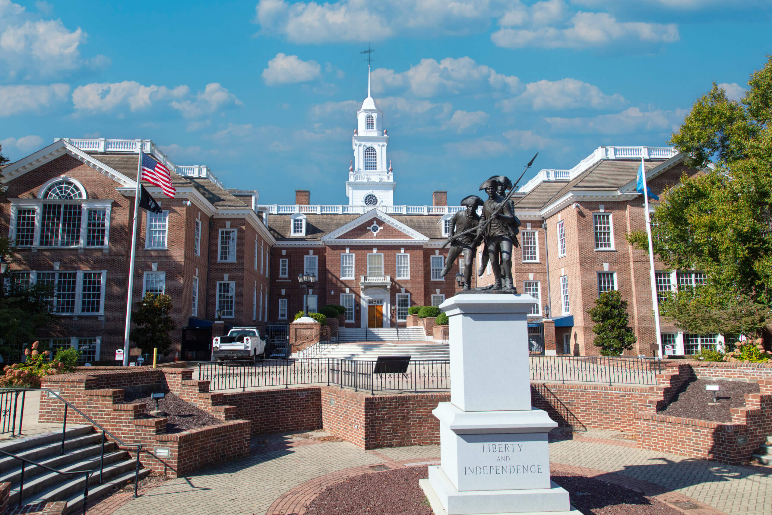 A brick government building with a clock tower, two flags, steps, and a statue in front labeled “Liberty and Independence” stands under a blue sky with scattered clouds—a fitting backdrop as Delaware 2026 business entity law amendments promise to shape the future within its historic halls.