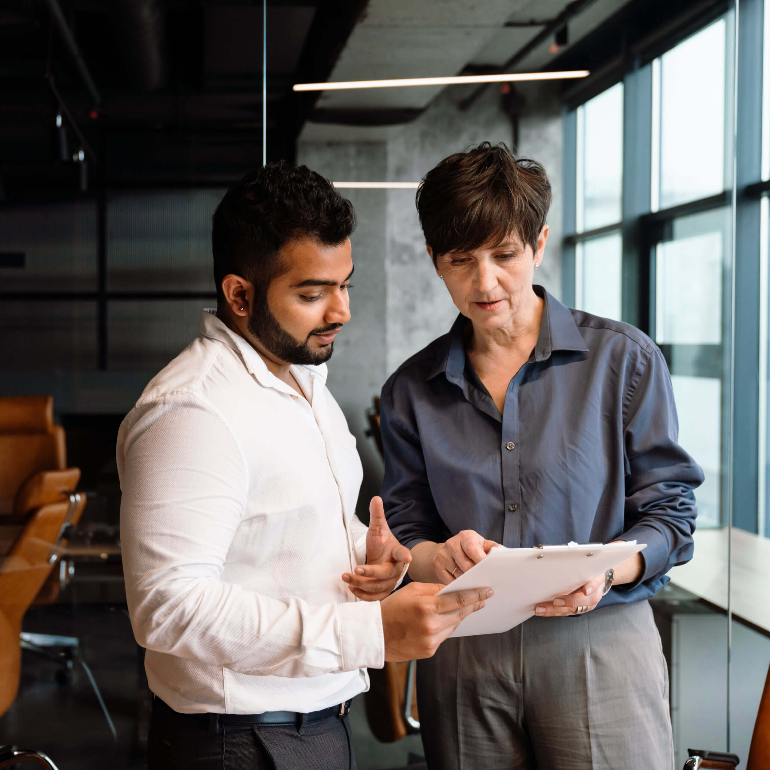 Two people stand together in a modern office, looking at and discussing a document on a clipboard. One gestures while the other holds the paper, both appearing focused and engaged in conversation.