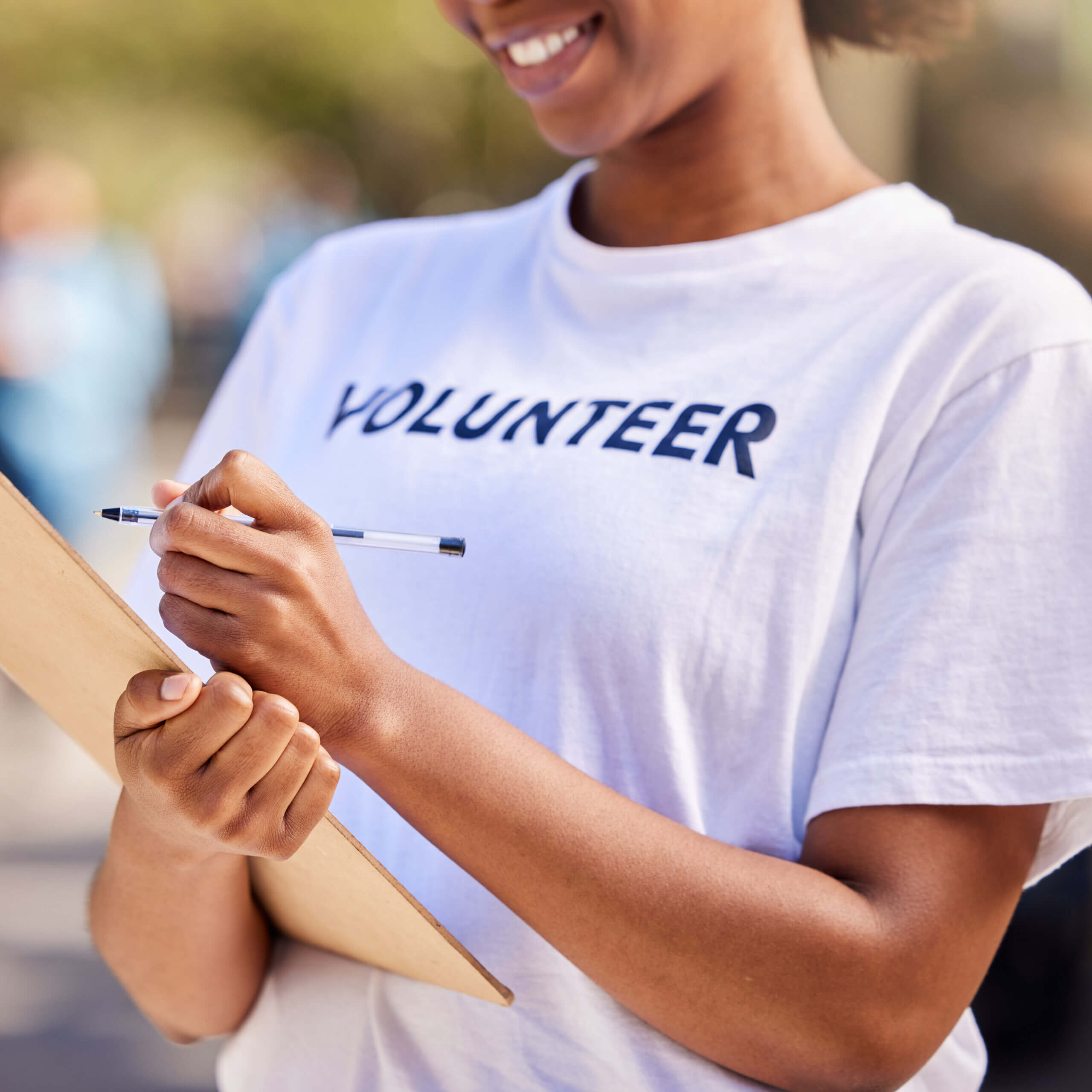 A smiling person wearing a white "VOLUNTEER" T-shirt holds a clipboard and pen, appearing to write or take notes outdoors. The background is blurred.