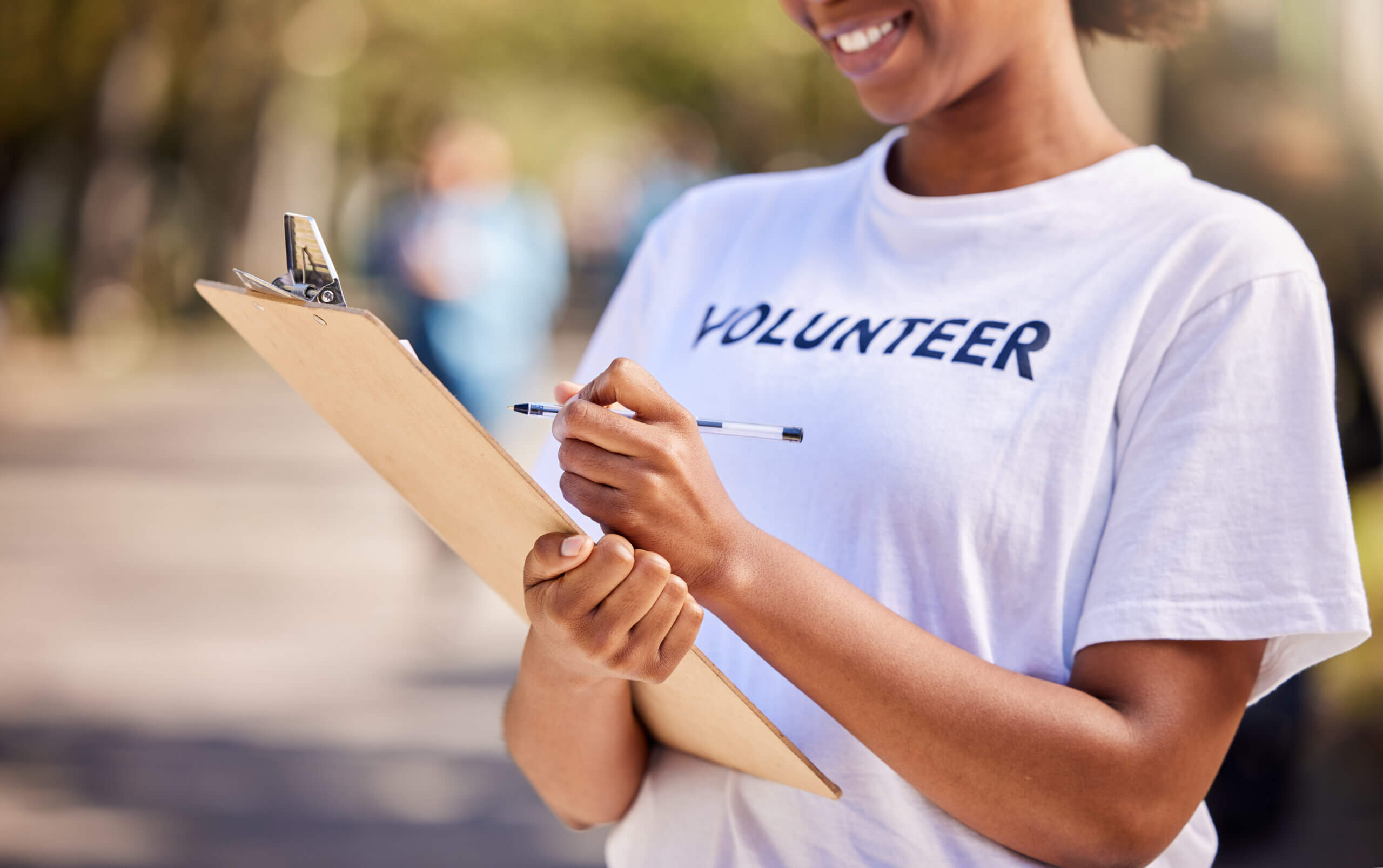 A person wearing a white "VOLUNTEER" t-shirt smiles while holding a clipboard and pen, likely filling out a form or survey outdoors. The background is blurred.