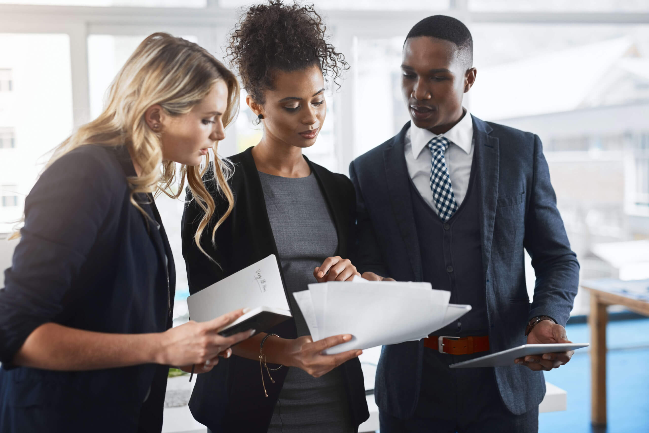 Three professionals stand together in an office, reviewing documents. One woman holds papers, while the others look on attentively. They are dressed in business attire and appear to be discussing work.