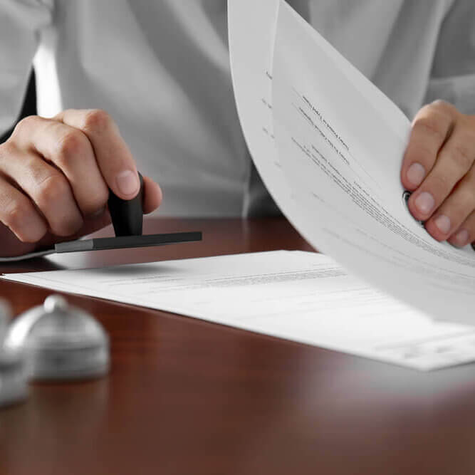 A person presses a stamp onto a document while holding several sheets of paper on a wooden desk. Other office items are slightly visible in the foreground.