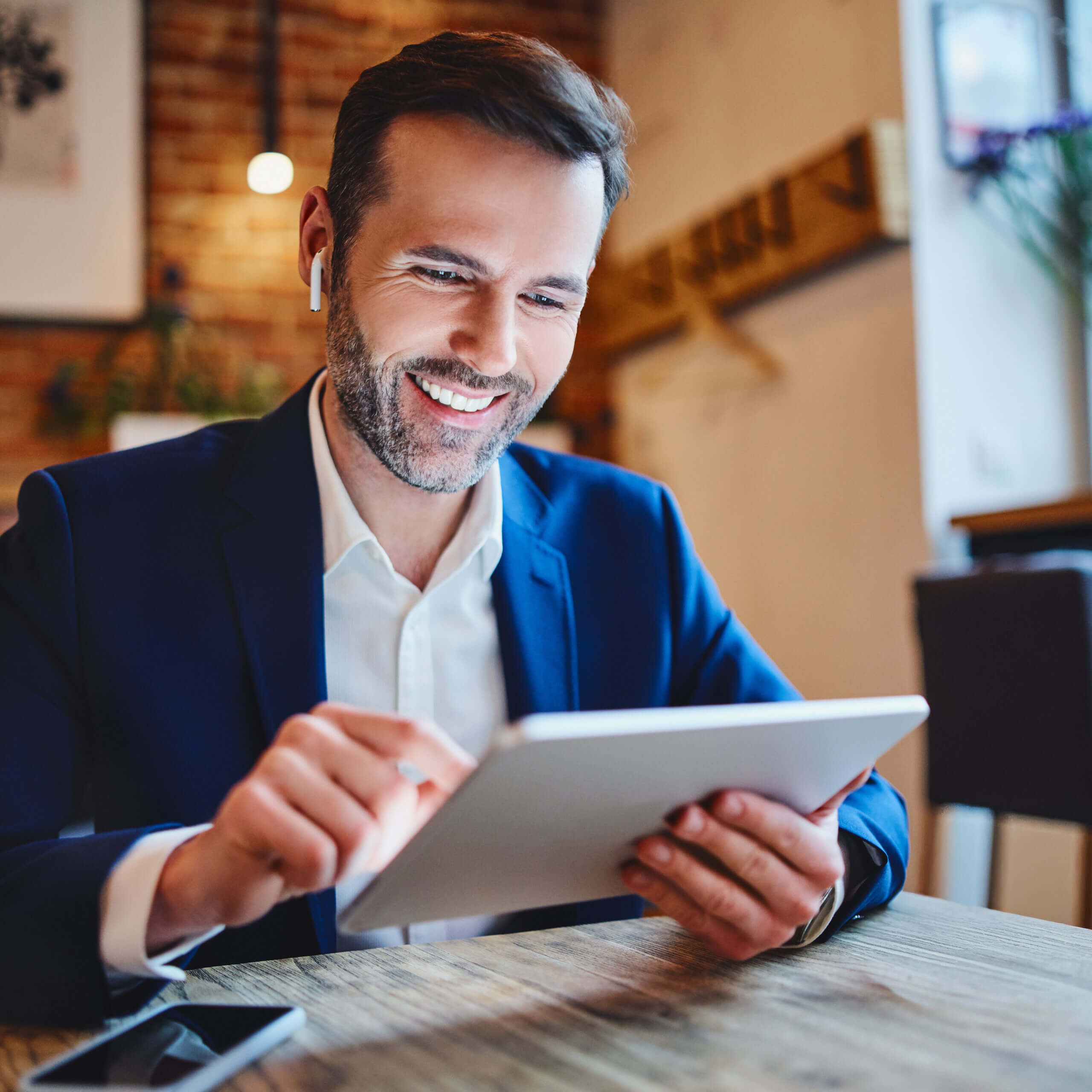 A smiling man in a suit sits at a wooden table using a tablet. He is wearing wireless earbuds and appears to be working or video chatting in a warmly lit, modern cafe.