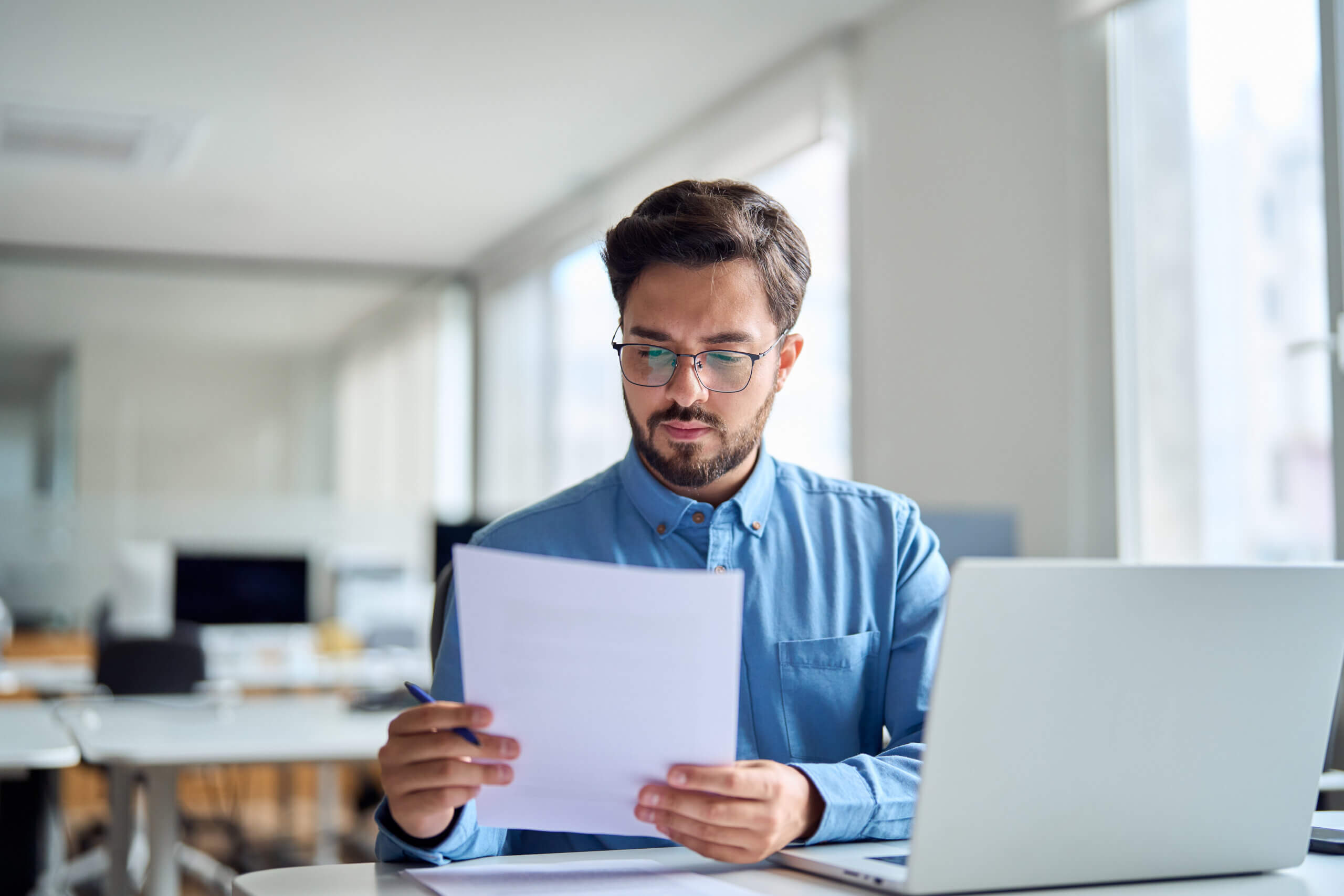 A man with glasses and a beard sits at a desk in a bright, modern office, reviewing documents related to a UCC debtor name. An open laptop and pen complete his focused workspace.