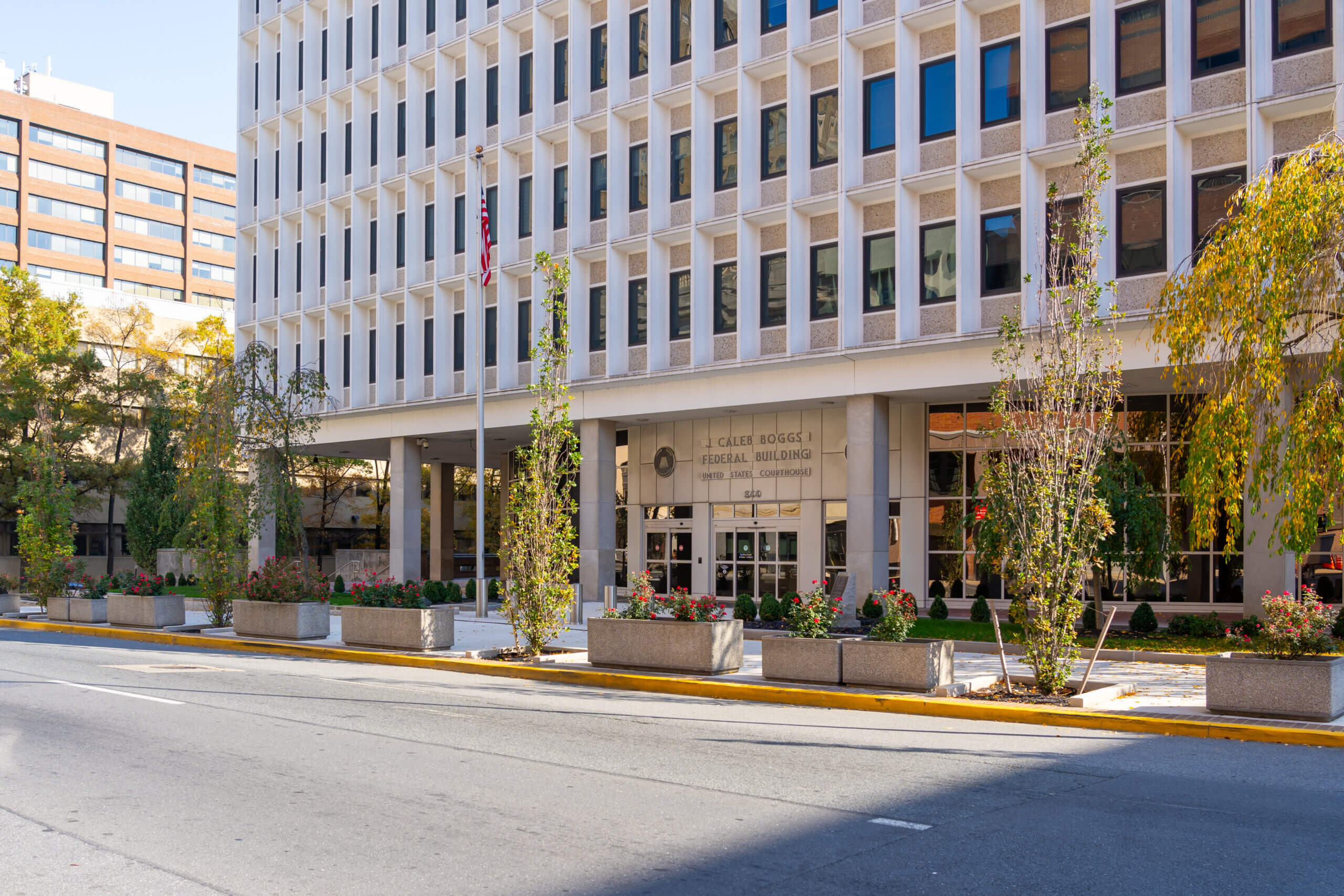 A multi-story white federal building with rectangular windows, labeled “J. Caleb Boggs Federal Building,” stands behind planters with young trees and flowers along a quiet city street—an important site for Delaware DBA filing services.