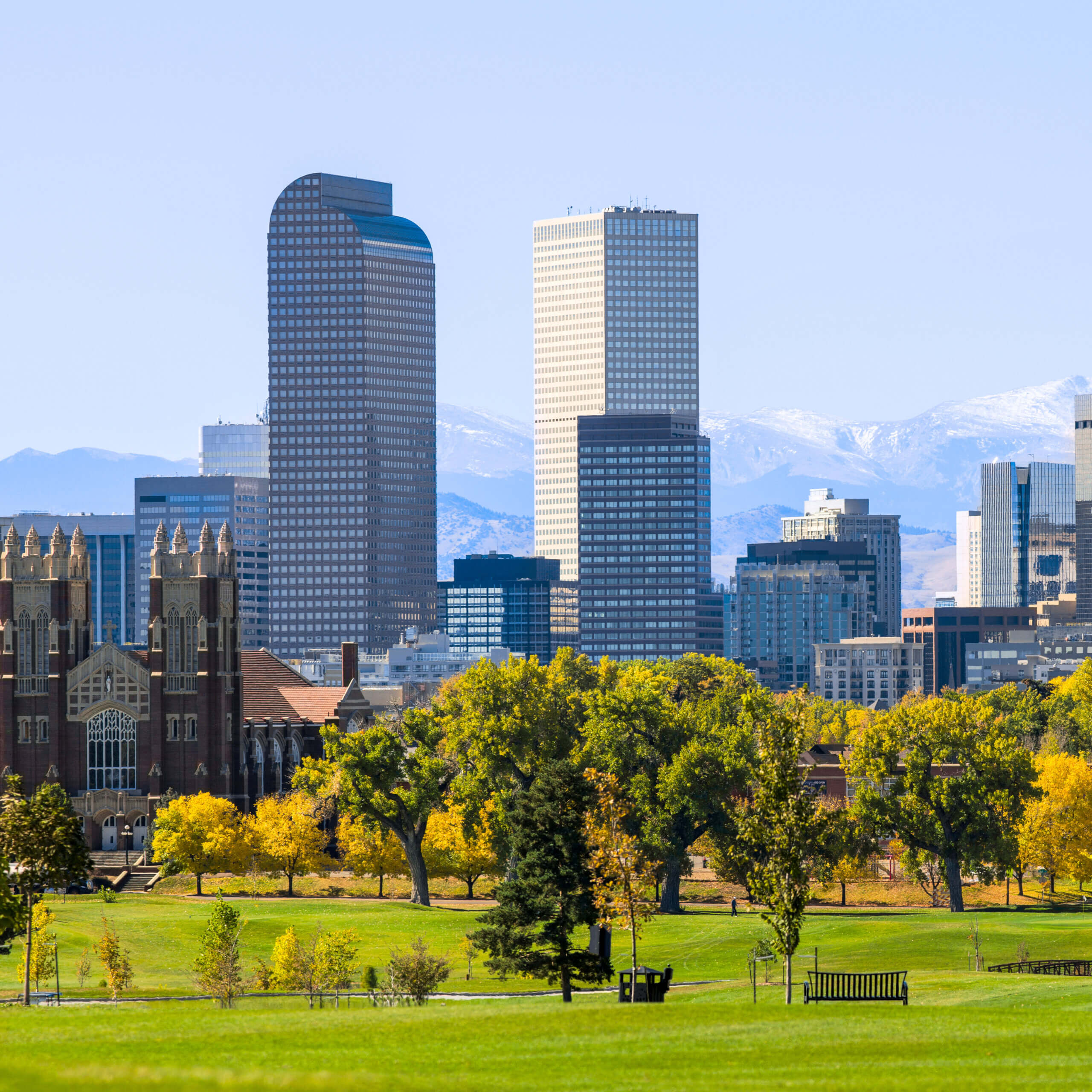 Denver skyline with tall modern buildings, a historic brick building, and green trees in the foreground. Snow-capped mountains are visible in the background under a clear blue sky.