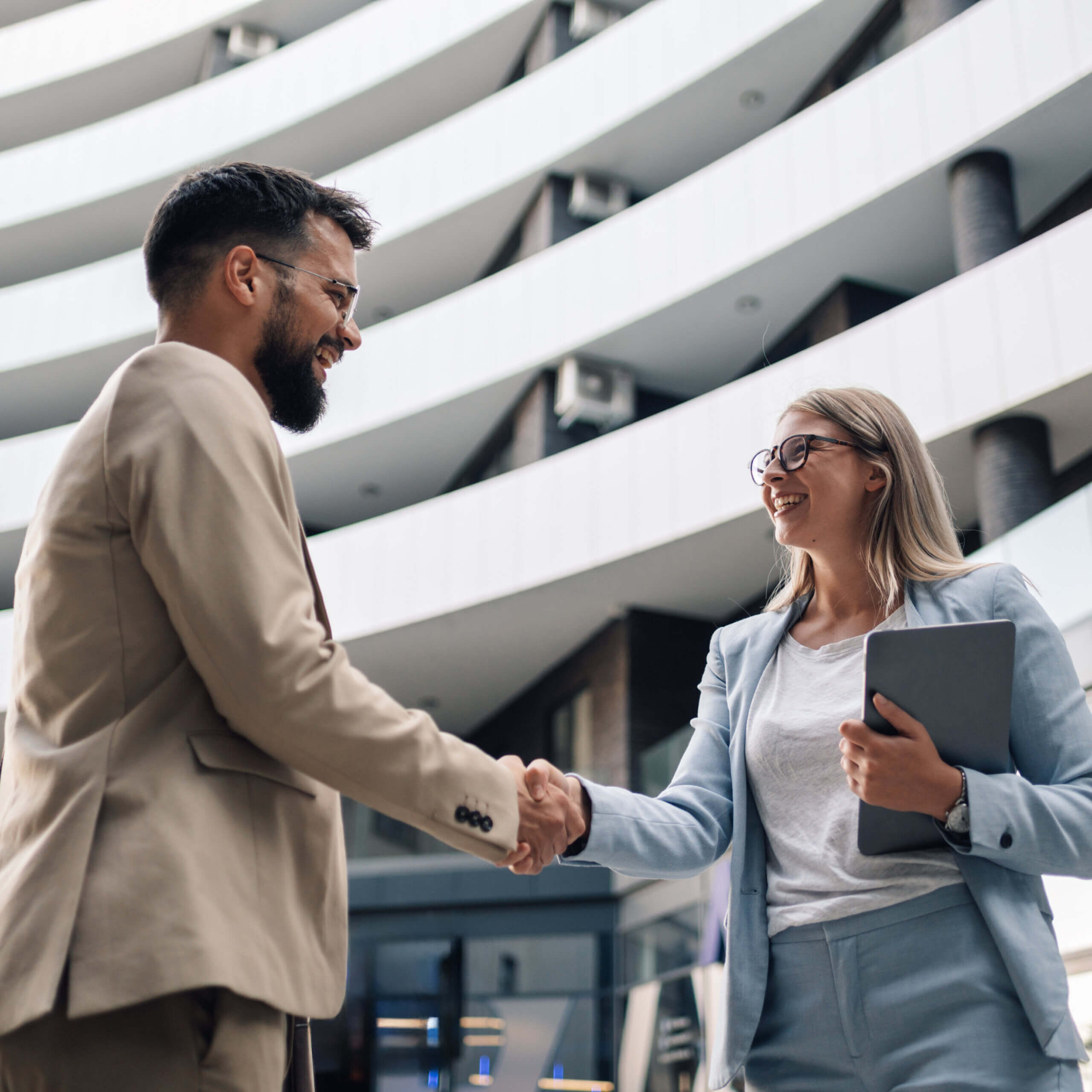 A man and woman in business attire smile and shake hands outside a modern office building. The woman holds a tablet and both appear to be having a positive, professional interaction.