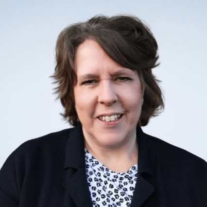 A middle-aged woman with short, wavy brown hair smiles at the camera. She is wearing a black jacket over a white shirt with a black floral pattern, appearing confident and approachable—perfect for discussing tax debt relief. The background is plain and light-colored.