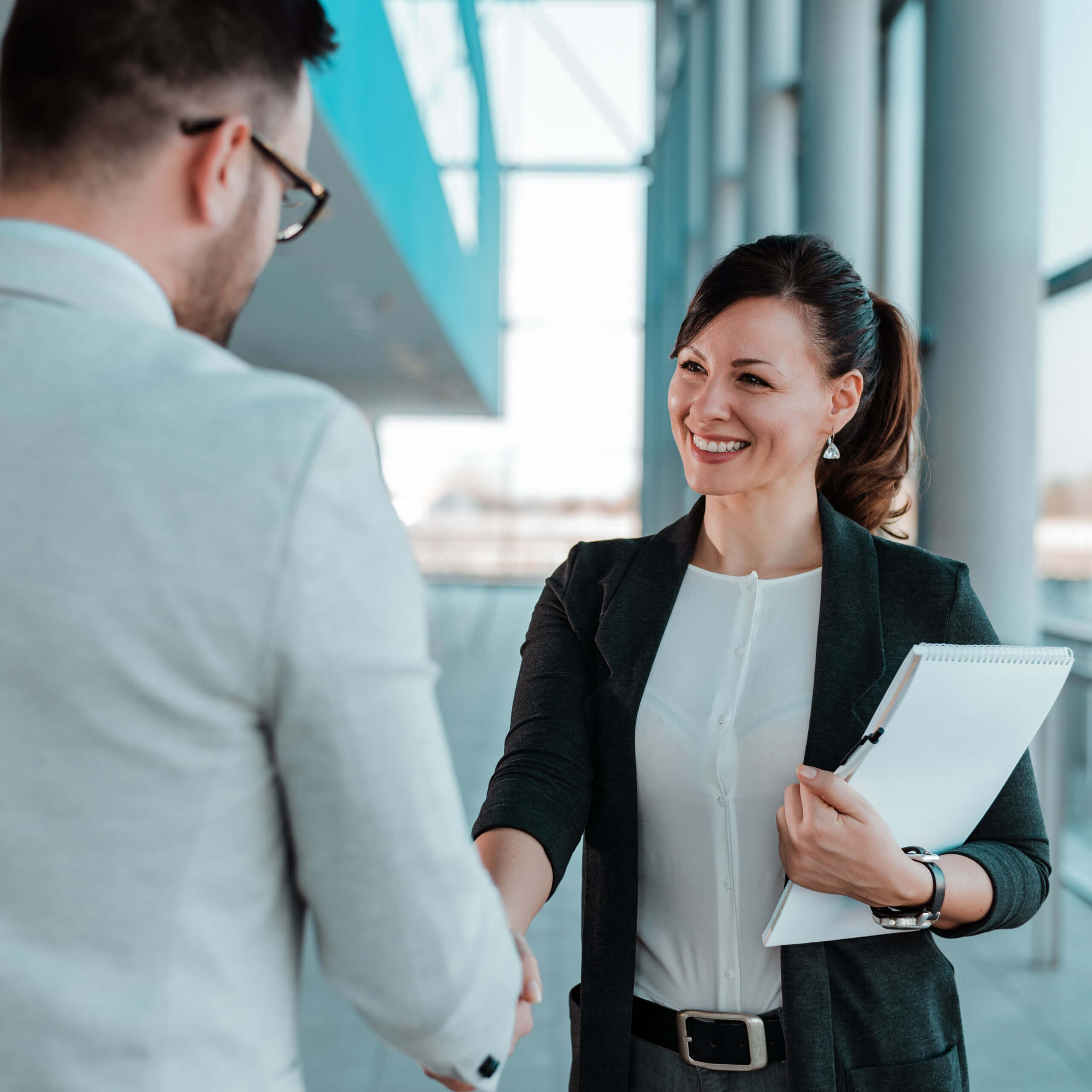 A woman in business attire smiles while shaking hands with a man in an office setting. She holds a notepad and pen, and both appear to be engaged in a professional interaction.