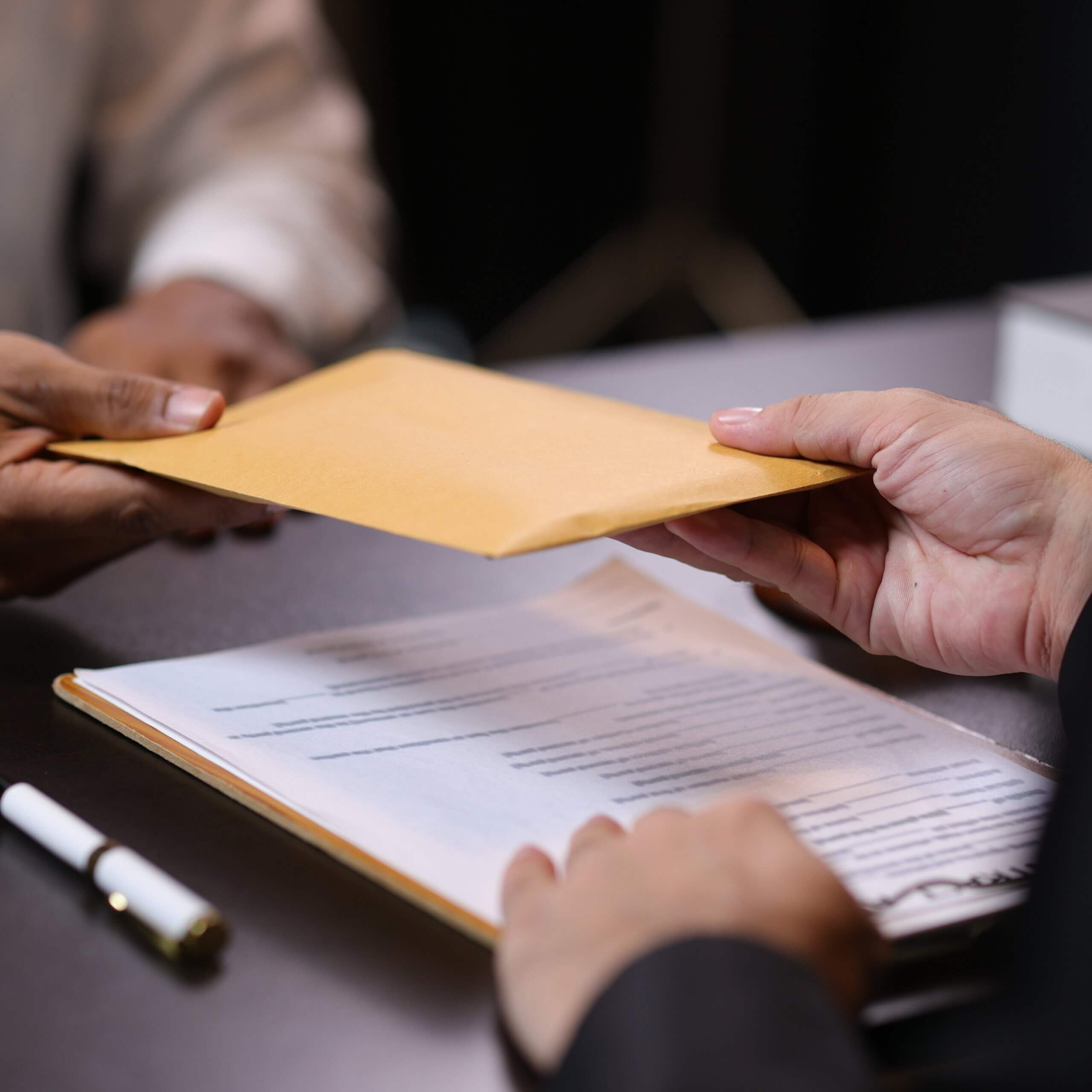 Two people exchange a large brown envelope over a desk with documents and a pen, suggesting a formal or business transaction.