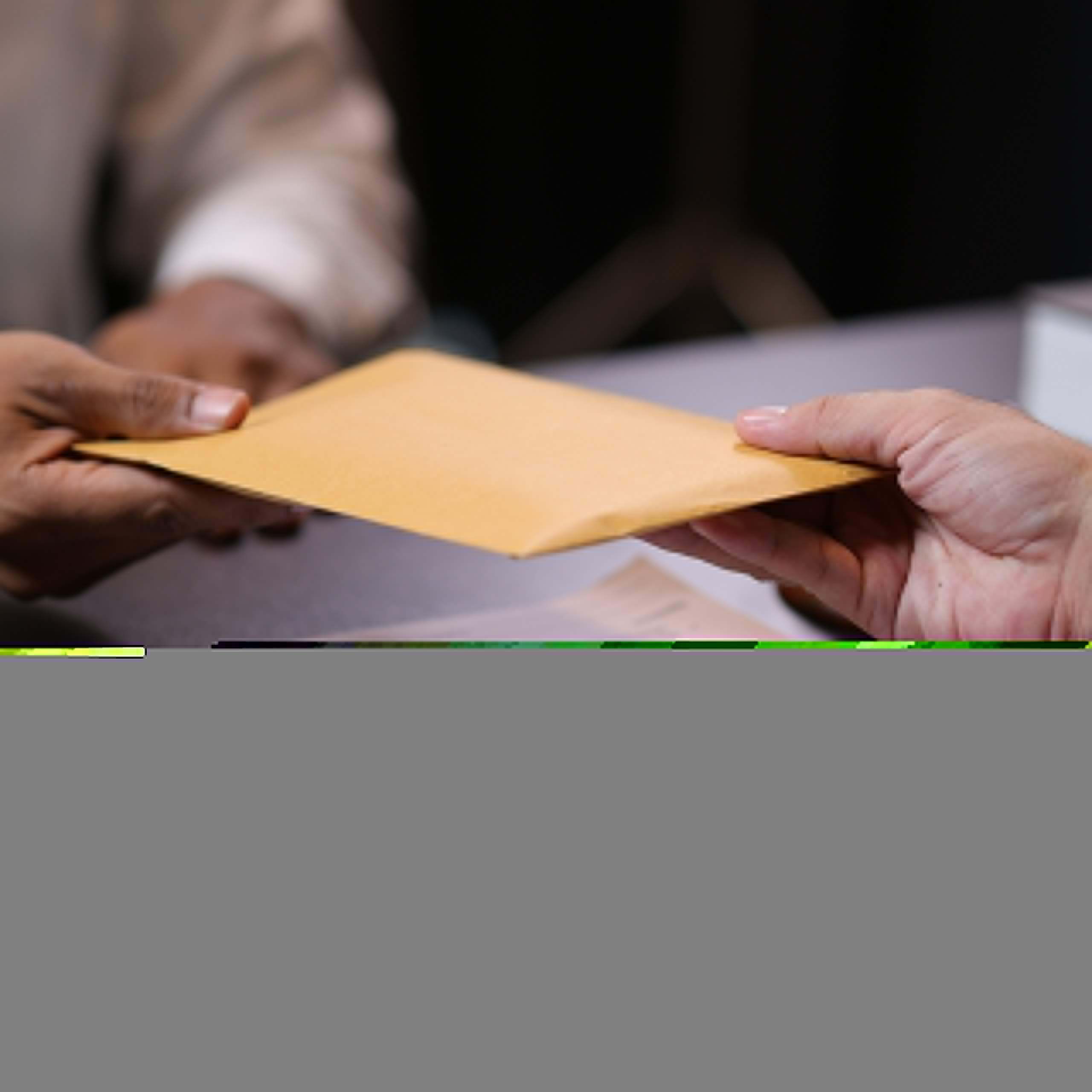 Two people exchange a large brown envelope over a table with documents and a pen, suggesting a formal or business-related transaction.