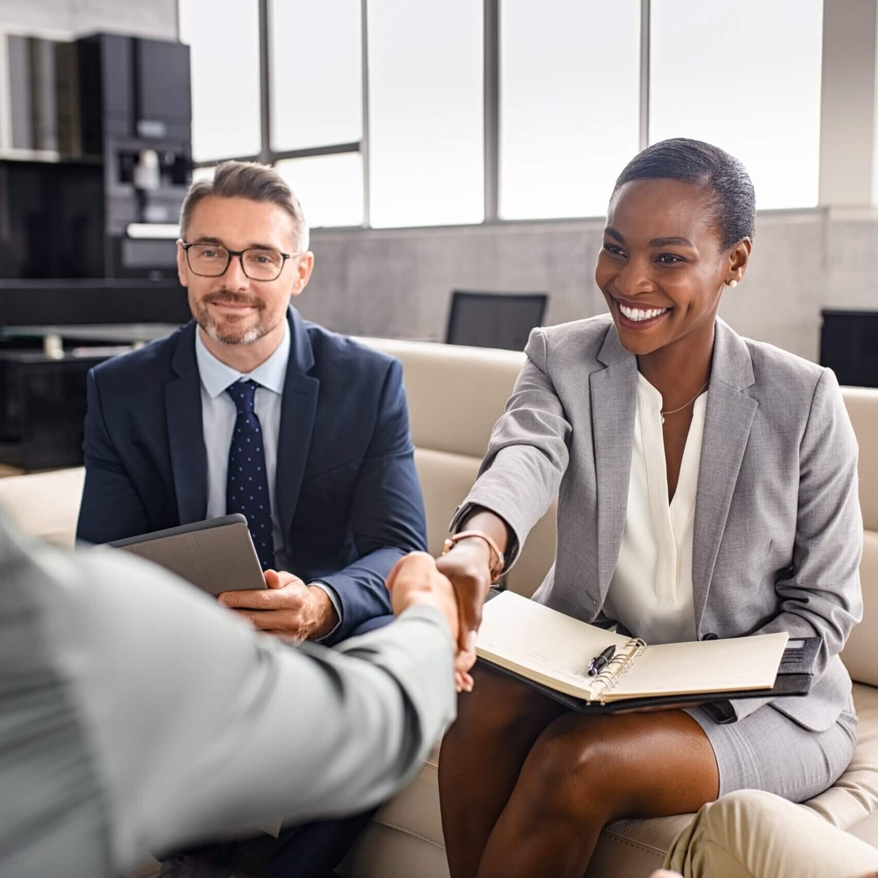 Two business professionals, a man and a woman in formal attire, sit on a couch during a meeting. The woman smiles and shakes hands with someone off camera, while the man looks on holding a tablet.