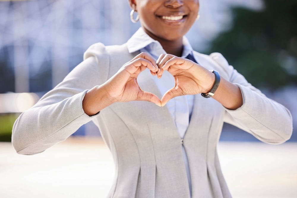 A person in business attire smiles while making a heart shape with their hands in front of their chest, symbolizing positivity and care. The background is blurred and outdoors.