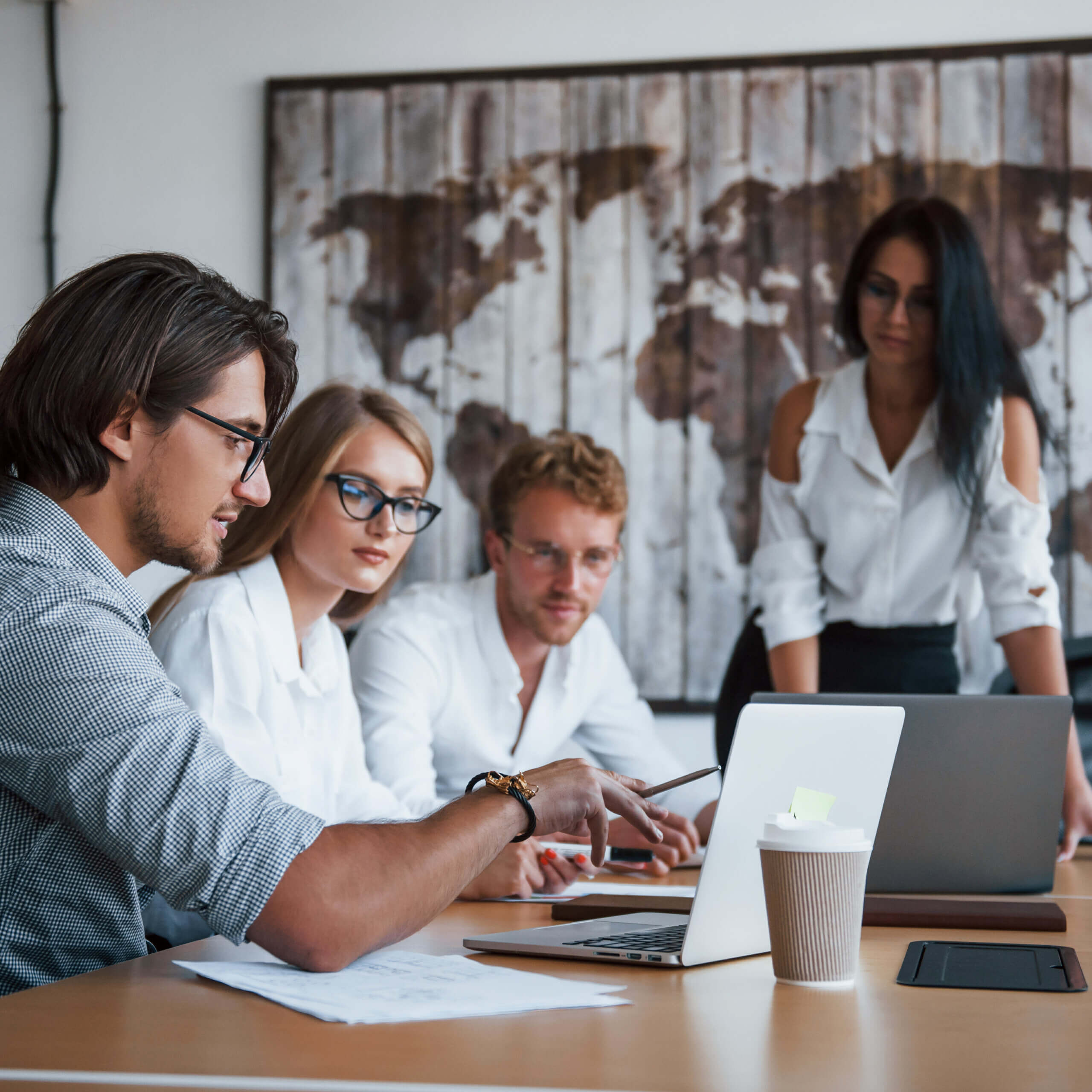 Four people in business attire sit and stand around a table with laptops, papers, and a coffee cup, engaged in discussion. A world map is displayed on the wall behind them.