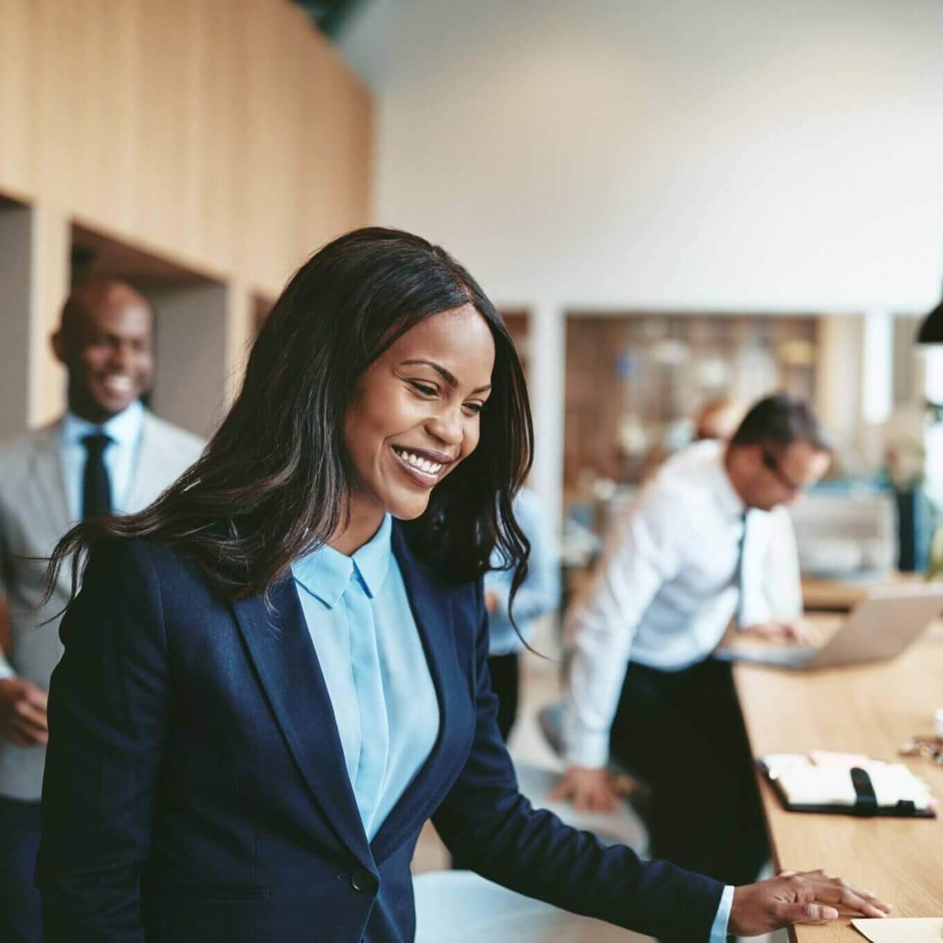 A woman in a blue suit smiles confidently in a modern office, with two colleagues working in the background.