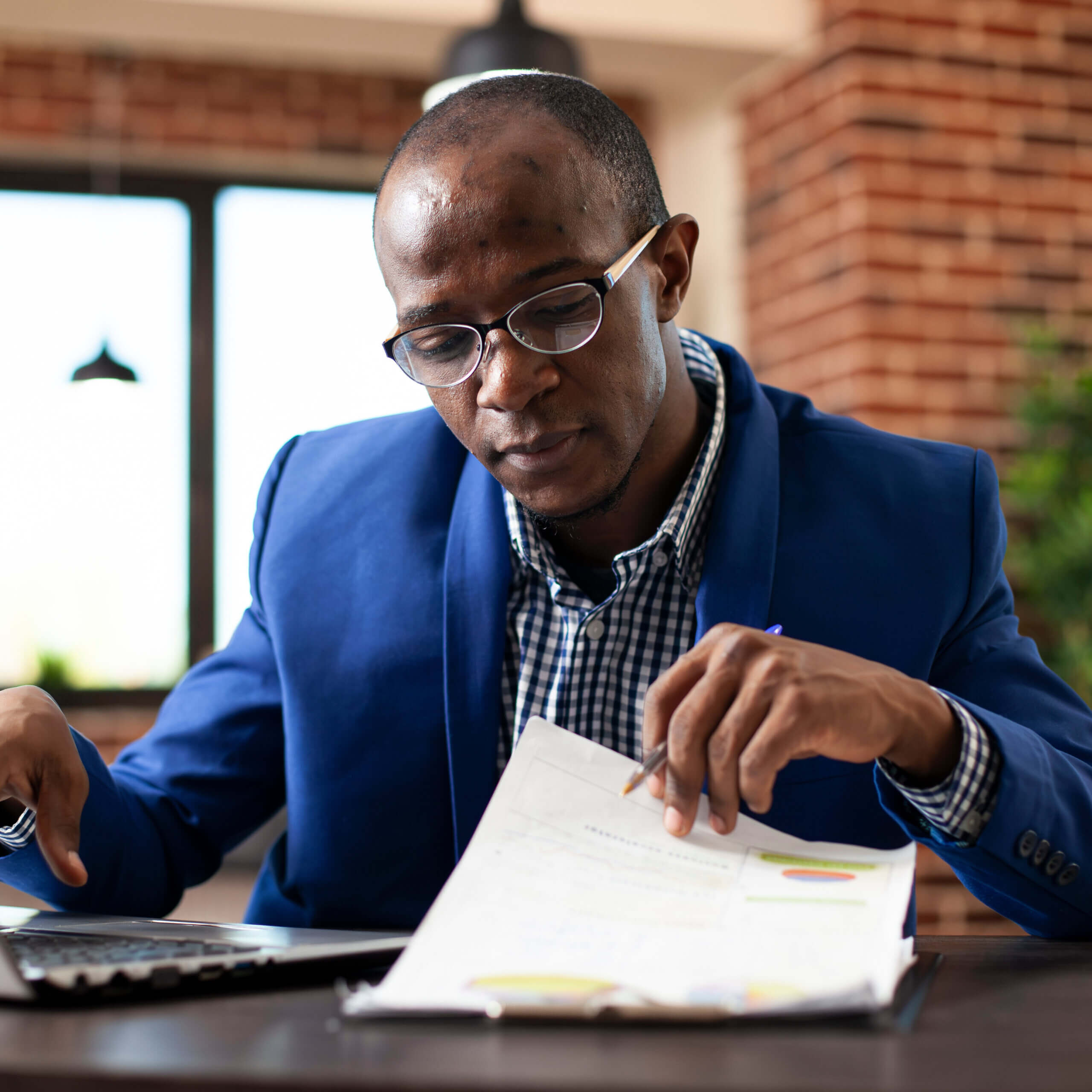 A man in a blue suit and glasses sits at a desk, reviewing papers with charts, while using a calculator. He appears focused, and the background shows a brick wall and a window with natural light.