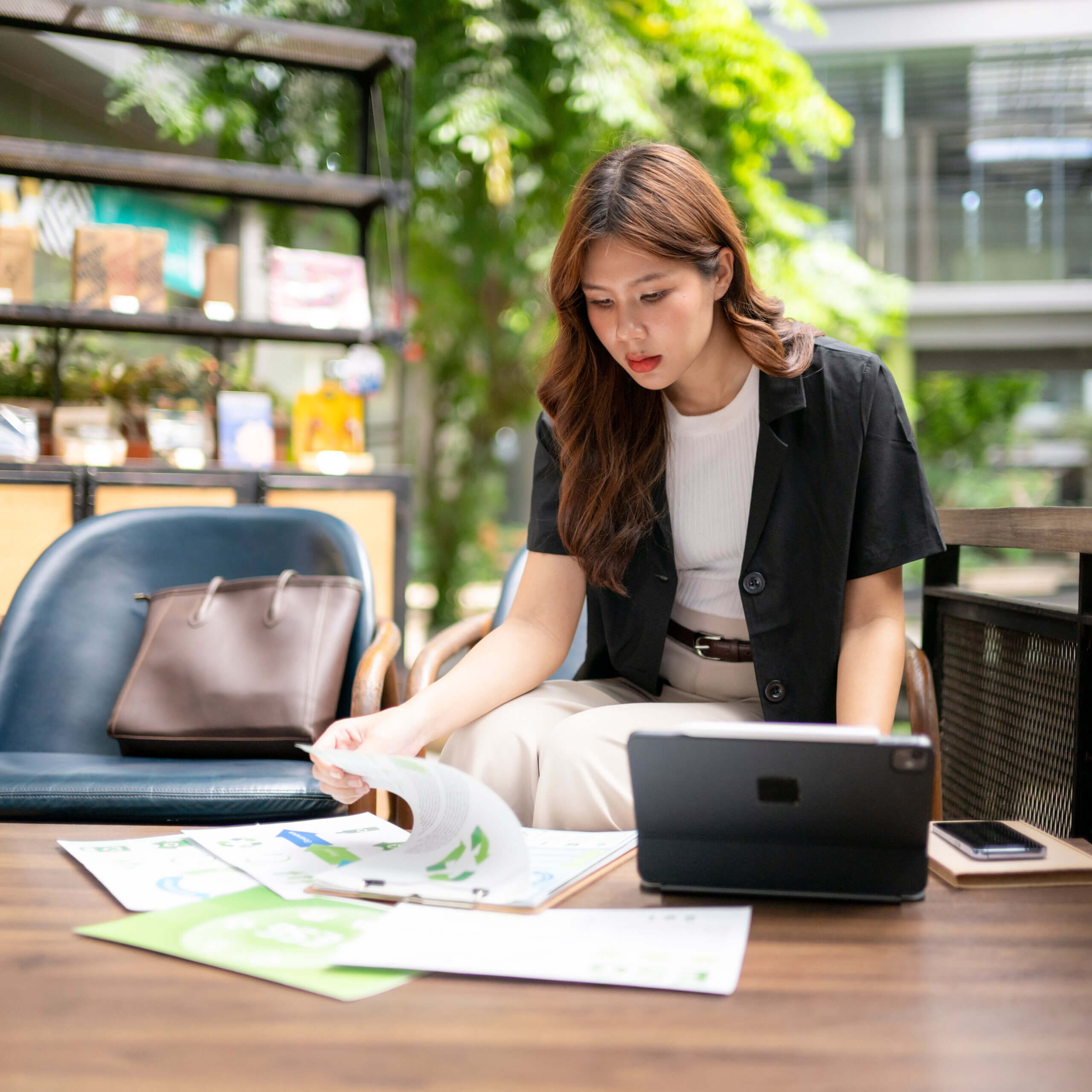 A woman sits at a table in a modern indoor space, reviewing documents and charts. A tablet, smartphone, and a brown bag are on the table beside her. She appears focused and engaged in her work.