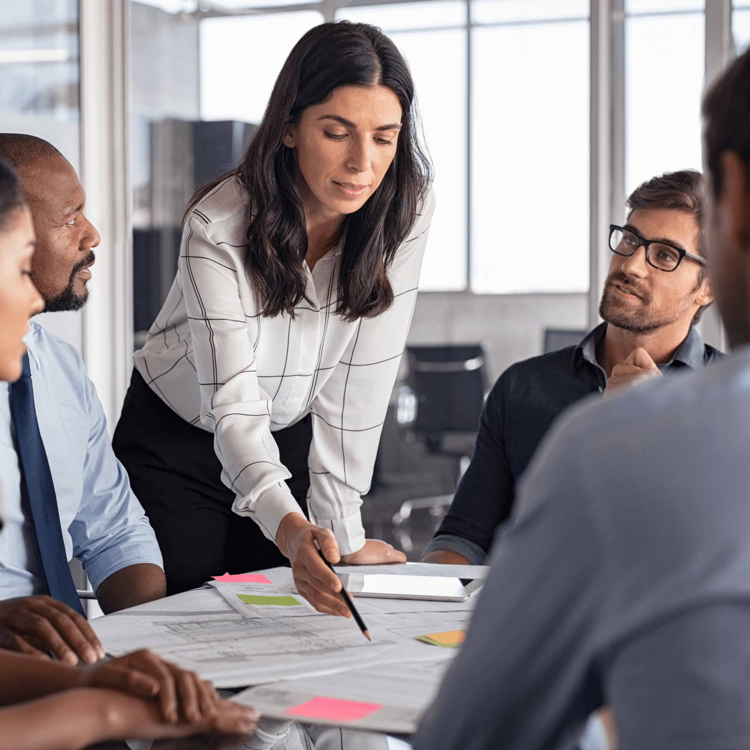 A woman stands and points at papers on a table while talking to four colleagues during a business meeting in a modern office.