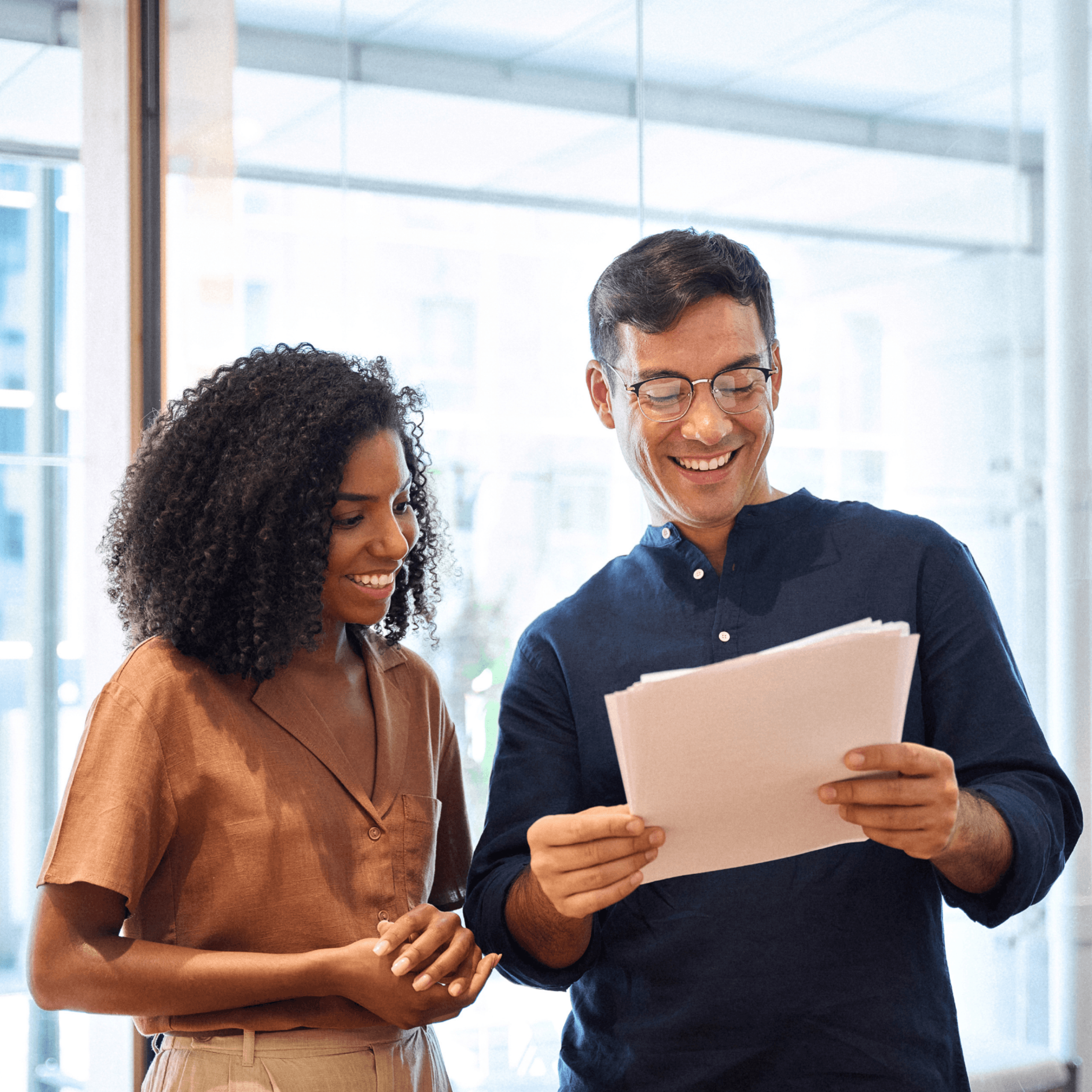 Two people standing indoors, smiling and looking at documents together in a bright office setting with large windows in the background—demonstrating seamless transaction support for law firms.