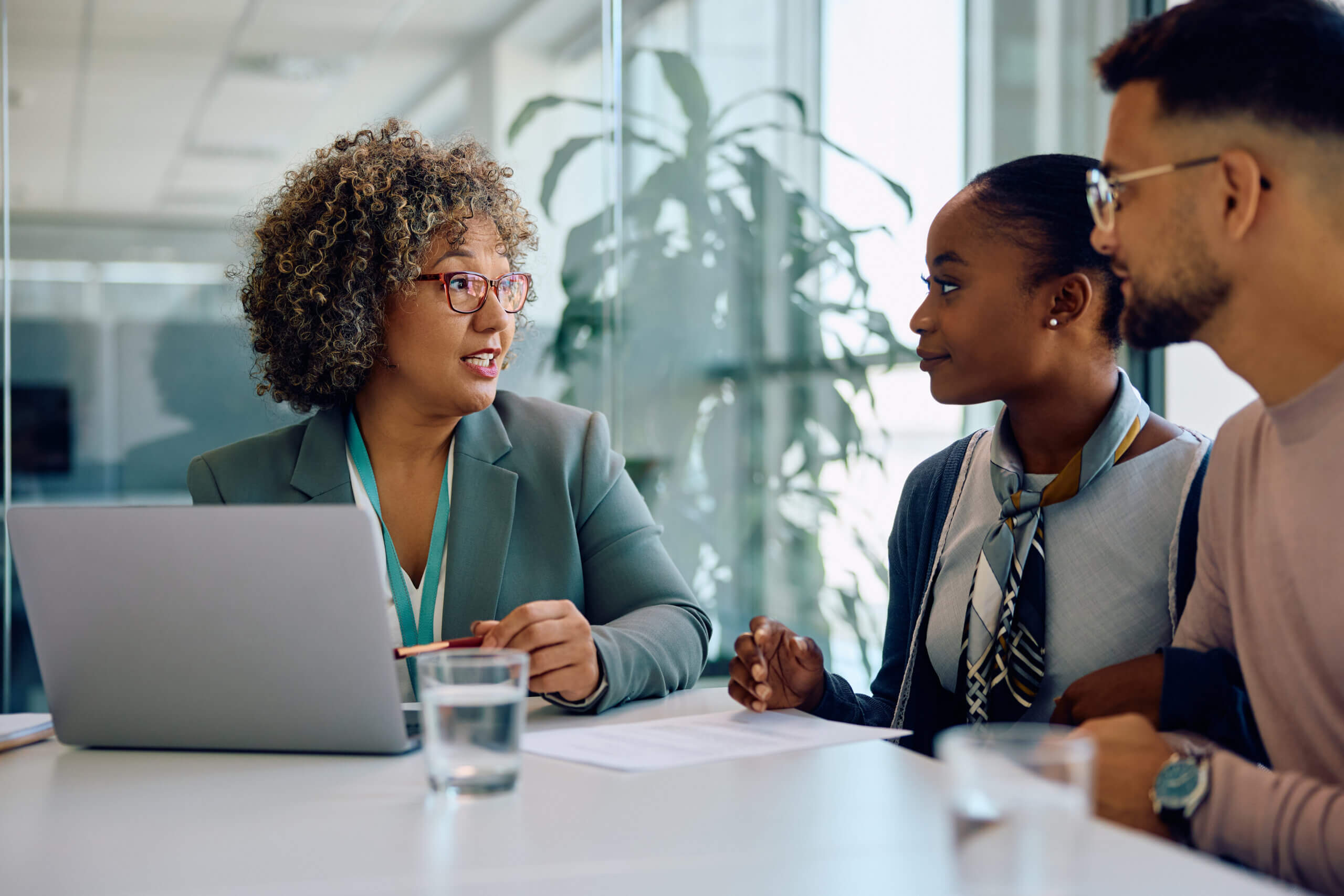 A businesswoman with curly hair and glasses discusses documents about process agent requirements with two colleagues at a table, featuring a laptop and water, in a bright, modern office setting.