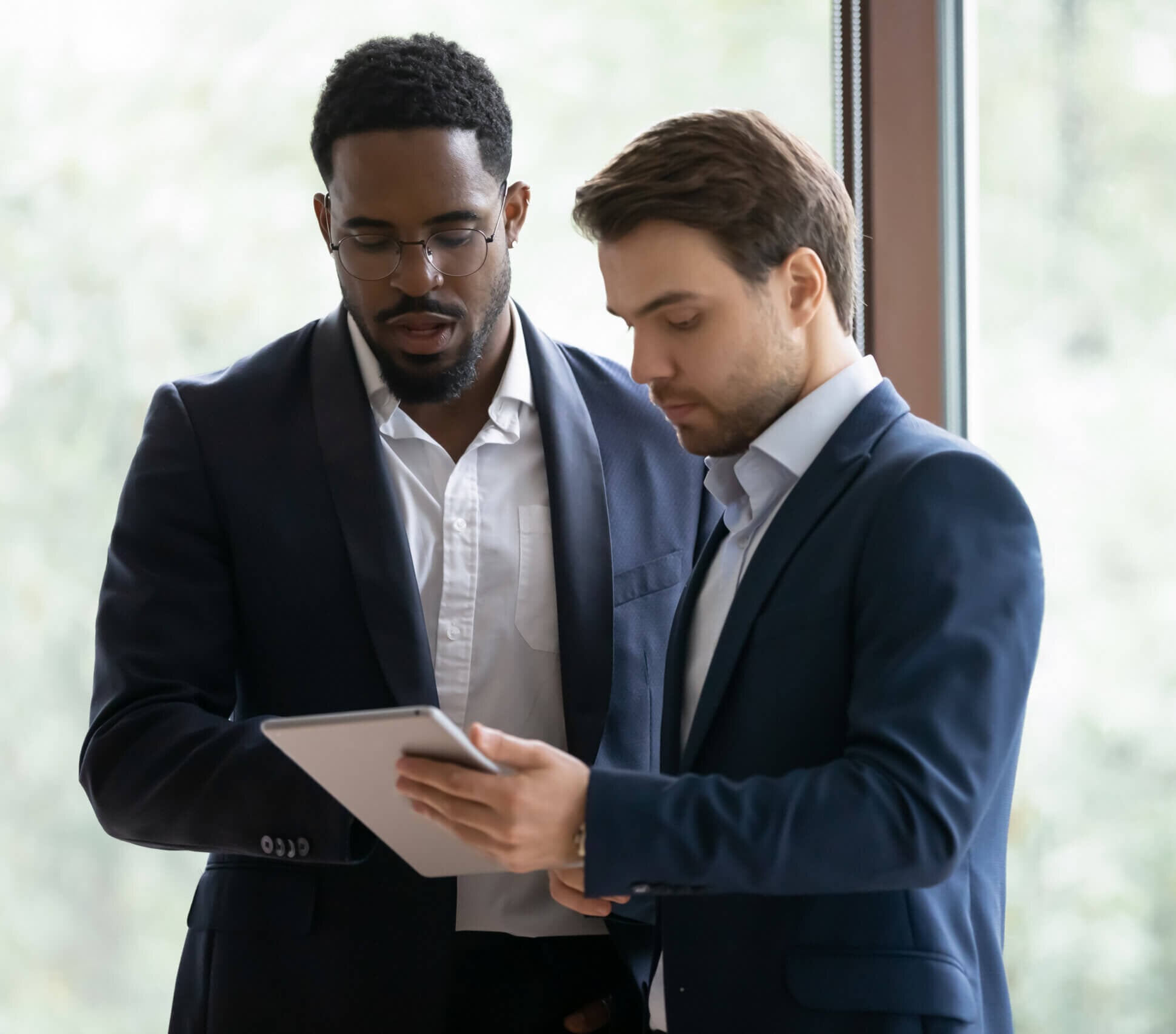 Two men in business suits stand indoors by a window, looking at a tablet together as they review International Due Diligence. One man holds the tablet while the other observes attentively. The background is softly blurred with natural light.