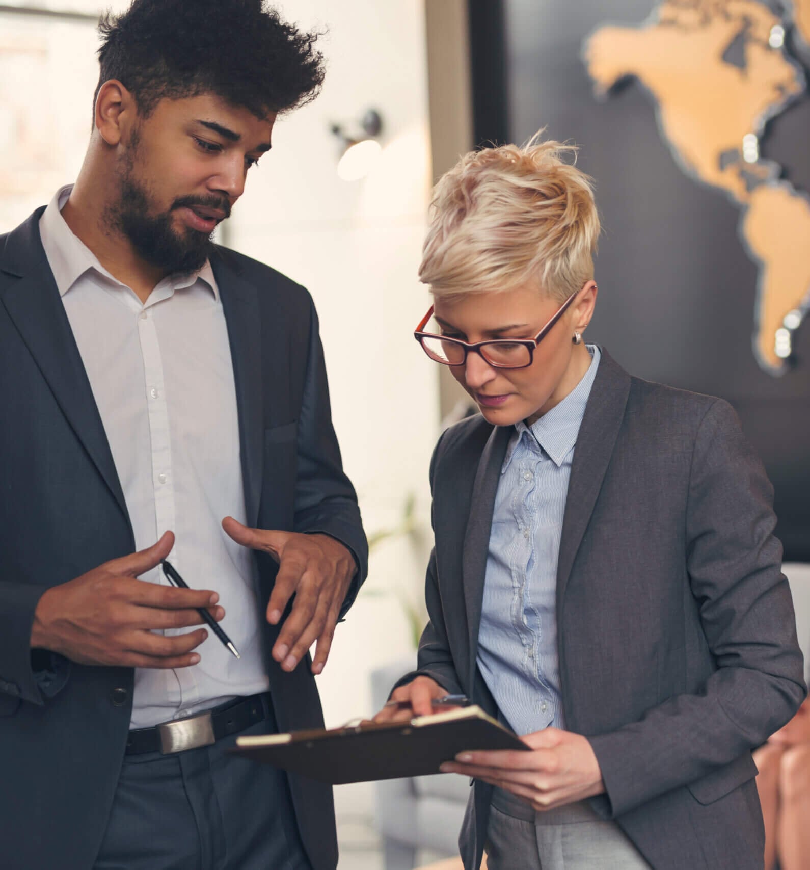 Two business professionals discuss a clipboard in an office, reviewing international due diligence documents, while four colleagues are seated and talking in the background. A large world map decorates the wall behind them.