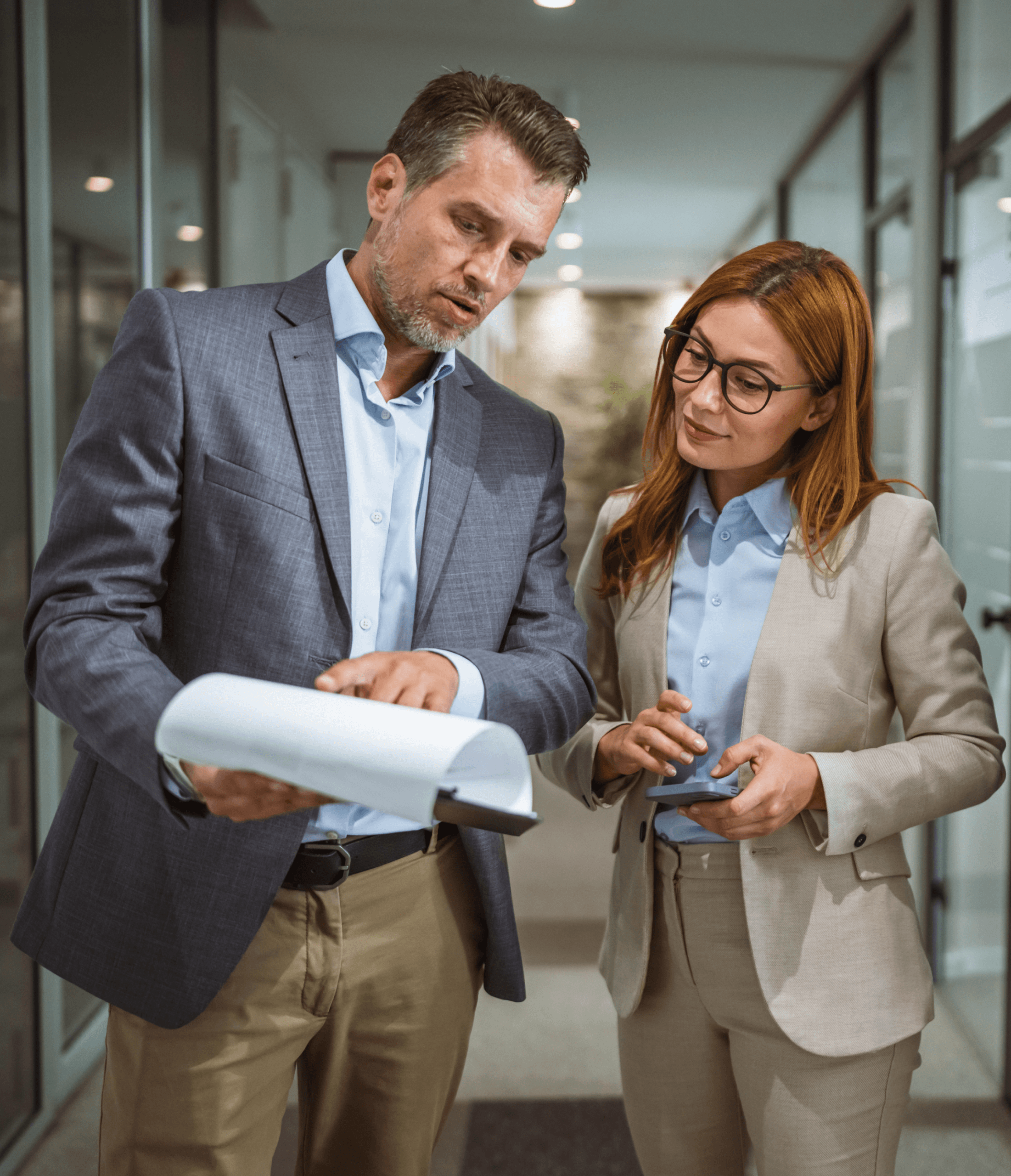 Two business professionals stand in an office hallway. The man in a gray suit points at papers, while the woman in a beige suit and glasses listens attentively, holding a tablet. They appear to be discussing work.
