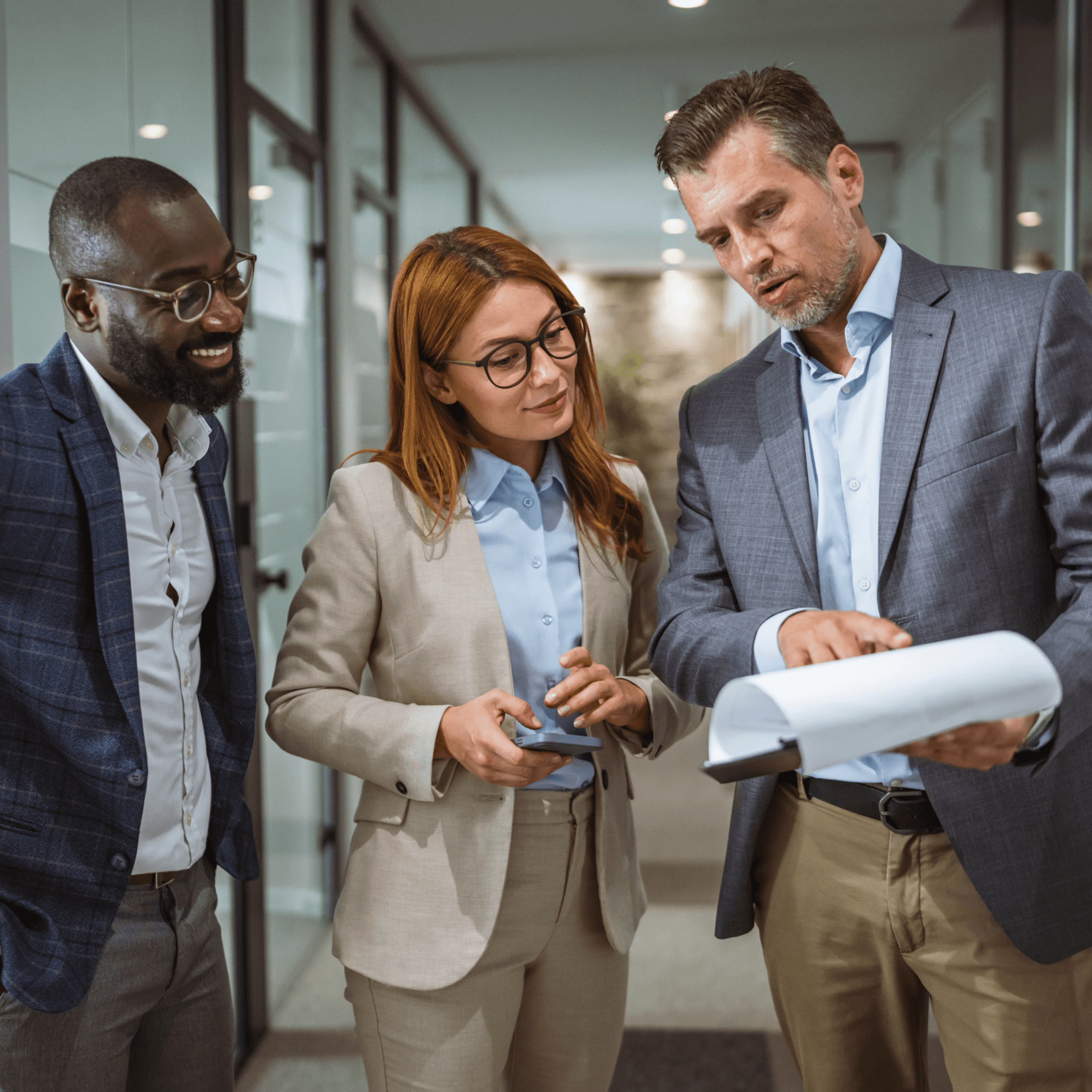 Three business professionals stand in a hallway, reviewing documents together. Two men and one woman, dressed in business attire, are engaged in discussion as they collaborate on law firm transaction support.