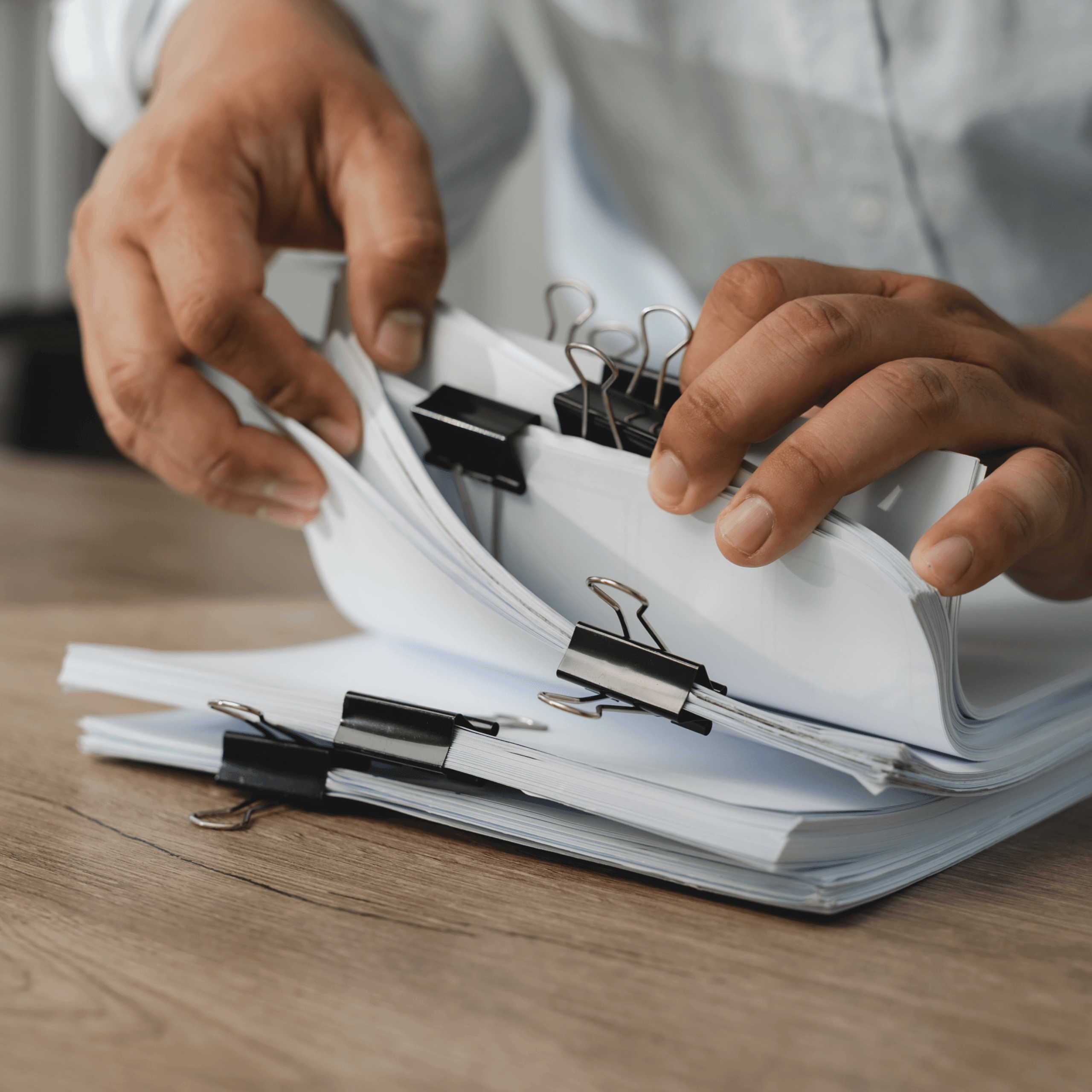A person organizes stacks of documents with black binder clips on a wooden table, wearing a light-colored shirt.